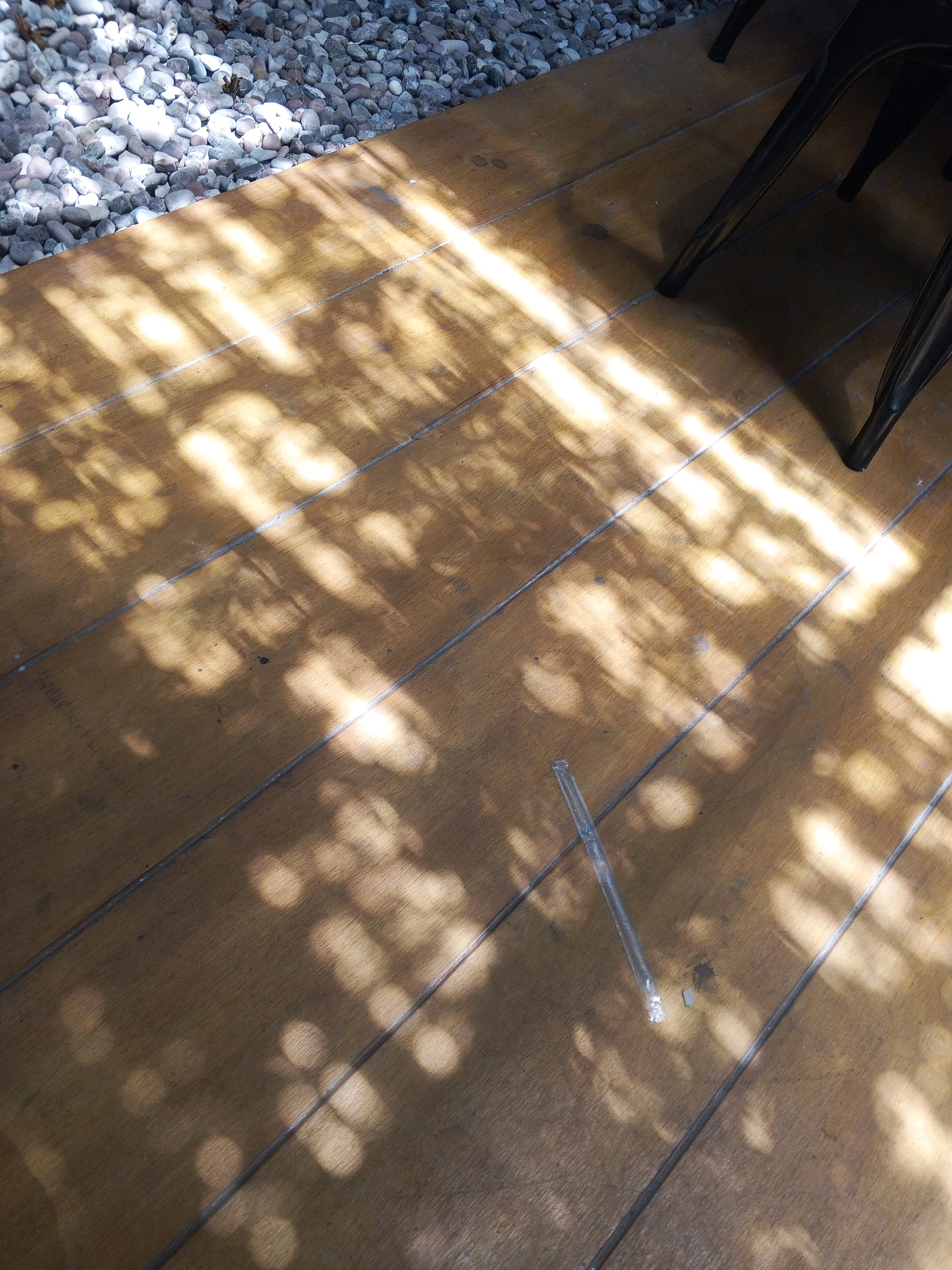 The image shows a close-up view of a wooden deck with dappled sunlight filtering through leaves, creating a pattern of light and shadow. The deck is composed of long, horizontal planks. In the upper left quadrant, there is a section of small, light-colored gravel. On the deck, a transparent plastic straw and a small, dark object are visible. In the upper right, parts of dark metal chair legs are present, suggesting an outdoor seating area. The scene is likely in Condongcatur, Indonesia, given the context.