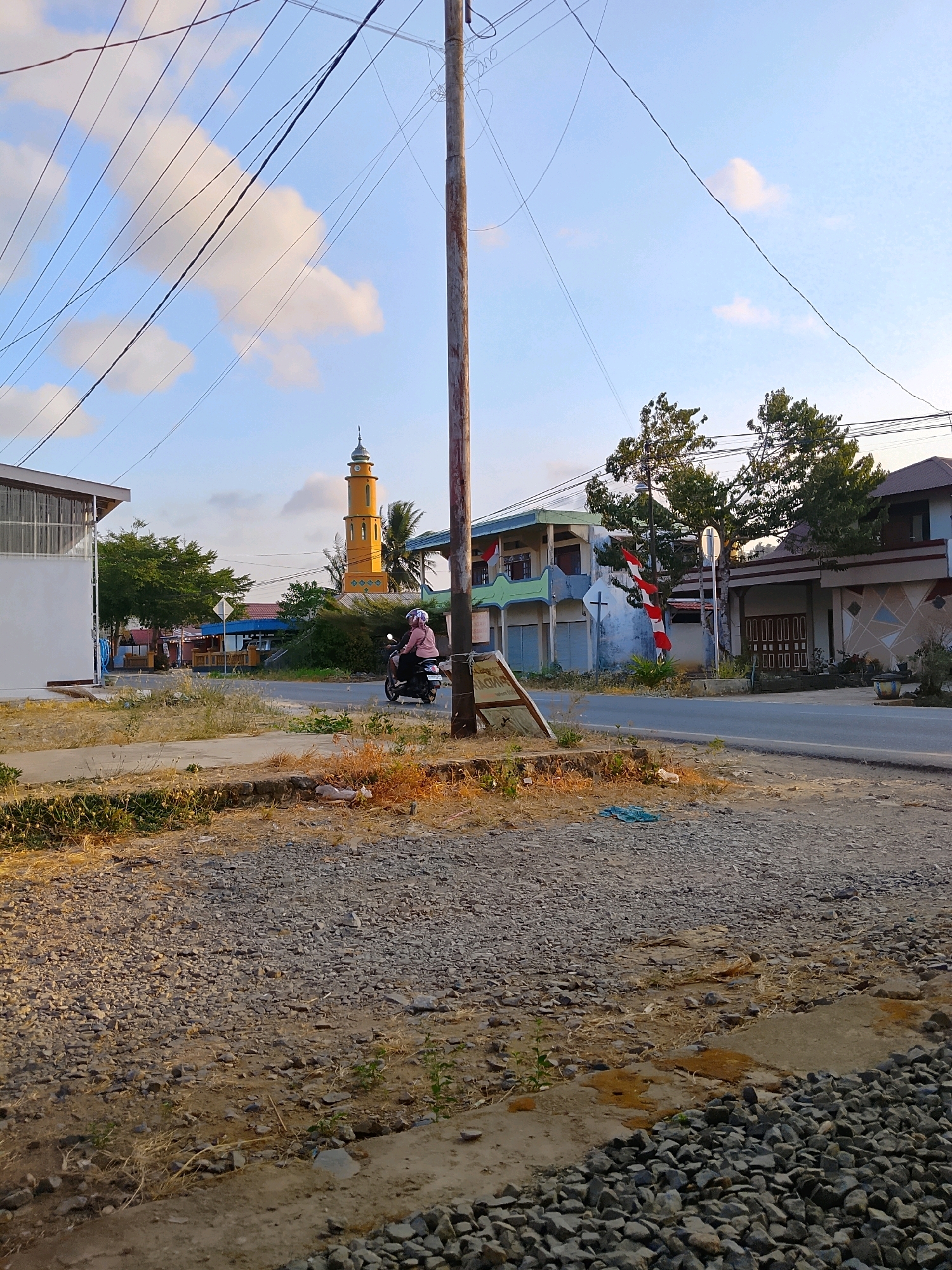 The image depicts a street scene in Pelaihari, Indonesia, characterized by a clear sky with scattered clouds and an array of overhead power lines. A tall, wooden utility pole stands prominently in the foreground, supporting the dense network of wires.
On the right side of the road, several buildings are visible. The closest building on the right has a light-colored facade with a patterned design and a dark, multi-paneled door. Next to it is a building with a green and white exterior, adorned with red and white Indonesian flags. Further down the street, a striking yellow minaret with a green-tipped dome rises above the other structures, indicating the presence of a mosque.
In the middle ground, a person rides a black scooter, wearing a pink top and a white helmet with a pink pattern. The scooter is moving away from the viewer on the paved road. The roadside is unpaved, covered with gravel and dry vegetation. Another building, predominantly white with a grey roof, is situated on the left side of the image, partially obscuring the view further down the street. The ground in the foreground is a mixture of gravel and soil, with patches of dry grass. The overall impression is that of a warm, daytime scene in a semi-urban or suburban area.