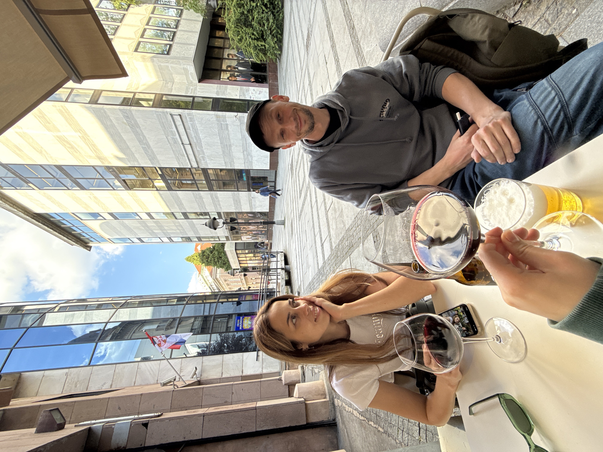 An outdoor, eye-level shot shows two people, a man and a woman, seated at a light-colored table at an outdoor cafe or restaurant patio in Belgrade, Serbia, on a sunny day. The perspective is slightly angled, with subjects looking towards the camera (man) or upward (woman).
The man, on the right, wears a grey hooded sweatshirt with a logo that appears to read "Truz" or "TRUE" and a black cap. He has a short beard and is smiling faintly, looking directly at the camera. He is wearing blue jeans and has a dark backpack resting on a chair behind him. His hands are clasped, holding a small black object.
The woman, on the left, has long brown hair and wears a white T-shirt with a gothic-style font text, partially visible, possibly "Trinity." She is smiling subtly and looking upward and to the left, with one hand gently touching her cheek. A third person's hand is visible on the far right, holding a wine glass.
On the table are two glasses of red wine, two glasses of beer with foamy heads, and a pair of green sunglasses. A smartphone, displaying what appears to be a group of people, lies face up between the wine glasses.
The setting features modern buildings with large glass windows reflecting the blue sky and clouds. In one reflection, a red, white, and blue flag, identifiable as the Serbian flag, is prominently displayed, confirming the location context of Belgrade. The ground is paved with patterned stone. Bright sunlight casts strong shadows, indicating midday. A sign in the background behind the woman contains Cyrillic text, partially visible as "МИЛИОН". The overall atmosphere is relaxed and pleasant.