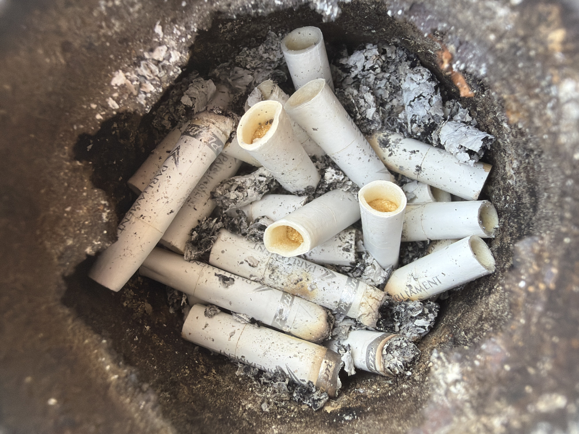 An overhead, close-up shot of a collection of cigarette butts fills the frame. The butts, primarily white with some yellowed filters, are densely packed within a dark, textured container. Ash and debris are scattered amongst the cigarette butts, some of which are partially burned and blackened. The overall impression is one of discarded waste.