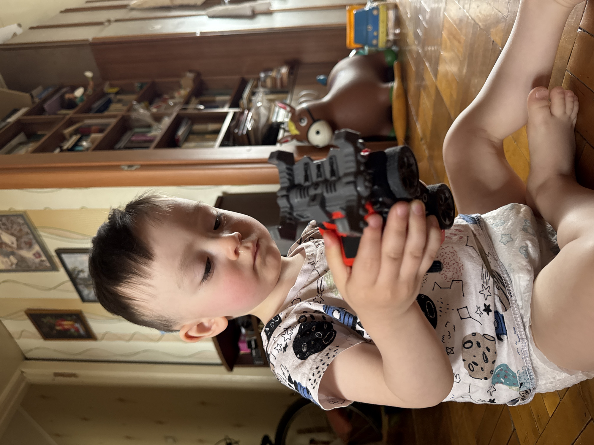 The image features a young child, likely a toddler, lying on a polished wooden floor and intently playing with a toy. The child is fair-skinned with dark, short hair and rosy cheeks, appearing calm and focused on the object in their hands. They are dressed in a light-colored t-shirt with a vibrant space-themed pattern, showcasing planets, stars, and rockets, along with a white disposable diaper or pull-up.
The toy the child holds is a dark grey and red vehicle, possibly an off-road car or monster truck, detailed with visible wheels and structural elements. The child's hands carefully grip the toy, and their gaze is directed downwards at it.
The setting is indoors, typical of a home environment. The child rests on a light brown wooden plank floor. In the background, a large wooden shelf unit or bookcase occupies a significant portion of the wall, filled with numerous books, various containers, and other household items. To the right, behind the child's toy, a distinct dark brown doll or figurine with prominent white eyes is visible on one of the shelves. Framed pictures or artwork adorn the wall to the left, above a subtly patterned wallpaper.
The scene portrays a peaceful moment of childhood play during what appears to be daytime, given the soft and even illumination. No visible text is present in the image. The context places this domestic scene in Izhevsk, Russia.