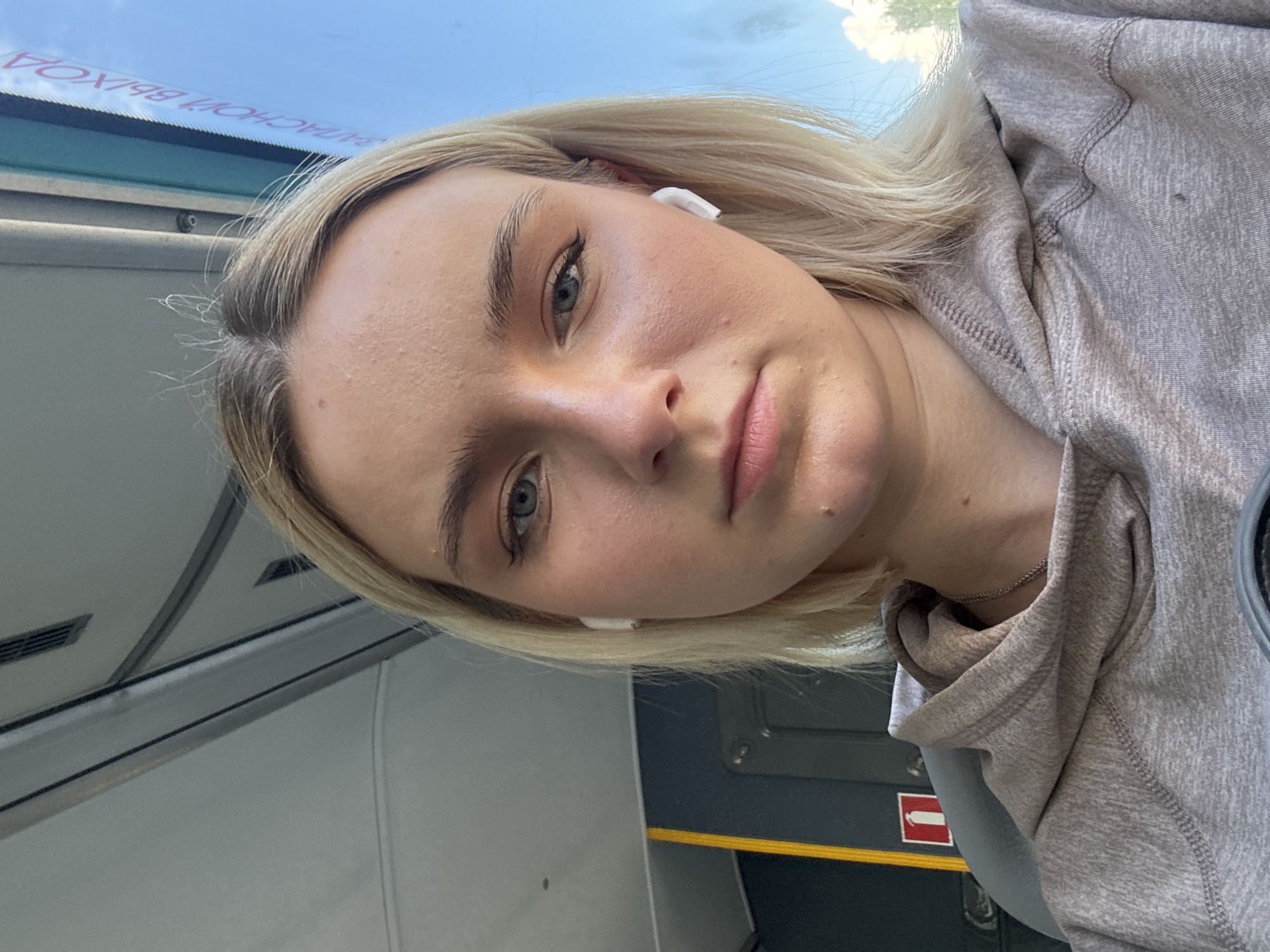 A close-up selfie captured from an upward angle shows a young woman with blonde hair and blue-grey eyes, wearing a light grey hooded sweatshirt. She has a neutral to slightly contemplative expression and is wearing white wireless earbuds in both ears. Her hair falls loosely around her face and shoulders.
The setting appears to be inside public transportation, likely a bus or train, given the overhead compartment-like structures, the window, and the visible signs. Through the window, a bright blue sky with some scattered white clouds is visible, suggesting daytime.
A significant detail is the inverted text visible through the window, which reads "ВЫХОД НА ОСТАНОВКЕ" (VYKHOD NA OSTANOVKE). This Russian phrase translates to "Exit at the stop" or "Stop exit," further indicating the location is a public transit vehicle. Another sign, partially visible in the lower right, shows a red rectangle with a white arrow pointing to the left, commonly used as an exit or direction indicator. The interior features grey panels, a yellow strip, and some dark grey elements.
The crops provide closer views of her face, showing her clear complexion, subtle makeup, and eyebrows, as well as the texture of her sweatshirt and the interior details of the vehicle. The overall impression is a casual portrait taken during travel, possibly in Novoe Devyatkino, Russia.