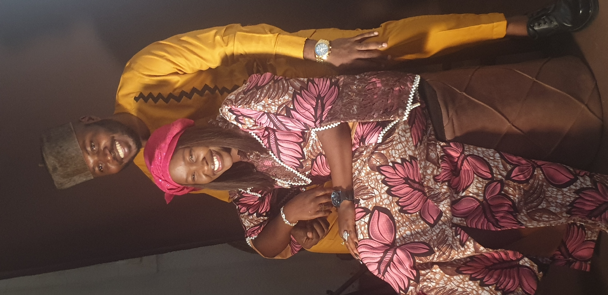 Two individuals, a man and a woman, are captured in a portrait, both smiling broadly at the camera. The image is taken indoors, with a dark, indistinct background, and a brown, quilted patterned object, possibly a couch or cushion, visible behind the woman. The overall lighting is dim.
The man, positioned behind the woman, wears a mustard yellow long-sleeved top, which features a black zigzag pattern across the chest. He has a short beard and is wearing a dark, possibly grey or brown, traditional cap. A prominent gold-colored watch with a metallic strap is visible on his left wrist. He is wearing black dress shoes, partially visible at the bottom right. His right arm is around the woman.
The woman is in the foreground, wearing a vibrant traditional African print dress, likely Ankara fabric. The dress features large, stylized pink and brown leaf or floral patterns on a light, mottled background. The neckline and sleeves of her dress are adorned with white beaded or lace trim. She wears a bright pink headscarf, and her long dark hair is visible beneath it. On her left wrist, she wears a dark-faced watch and a silver-colored bracelet. Rings are visible on her fingers. Her left hand is placed over the man's right hand.
Both individuals appear happy and joyful, with wide smiles, suggesting a celebratory or pleasant occasion. Their traditional attire and cheerful expressions contribute to a warm and inviting atmosphere. There is no visible text in the image.