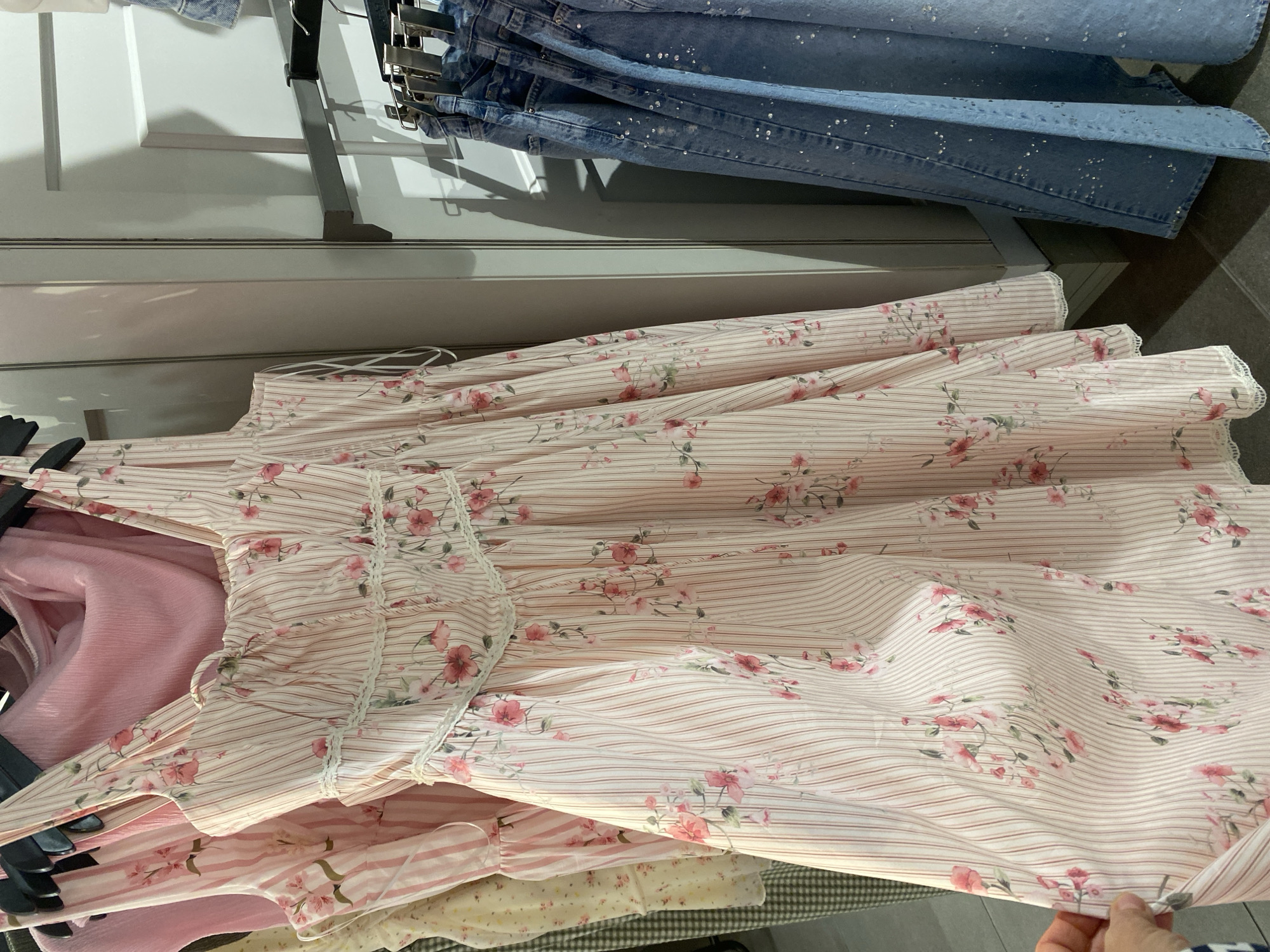 The image displays an indoor scene, likely within a clothing store or boutique in Alanya, Turkey, showcasing garments on display racks.
In the foreground, a person's hand holds up a light-colored dress featuring a distinct pattern. The dress fabric has thin, reddish-brown vertical stripes on a cream or pale pink background, adorned with scattered clusters of pink and green floral motifs, resembling roses or peonies. The bodice of the dress exhibits delicate lace trim and ruched details. Beneath this main garment, other clothing items are partially visible, including a plain pink textured fabric and other pieces with similar floral or striped patterns.
Above the floral dress, several pairs of light blue denim jeans are neatly hung on a metal clothing rack. These jeans are notable for their embellishment with numerous small, sparkling specks, which appear to be glitter or tiny sequins. The background shows white and grey wall or shelving panels, with shadows cast by the clothing rack, suggesting artificial indoor lighting. The scene presents a well-organized retail display of fashionable clothing. No visible text is present in the image.