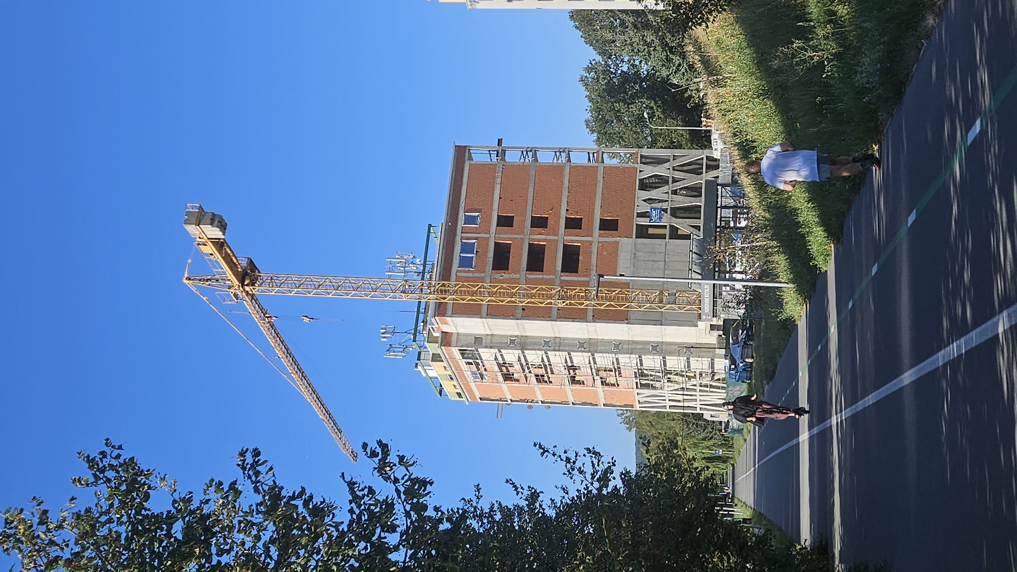 The image depicts a construction site in Prešov, Slovakia, on a clear day with a bright blue sky. A prominent yellow tower crane is positioned next to a multi-story building under construction, which is partially clad in red brick. The building has visible concrete structural elements and numerous open window frames. Atop the building, several communication antennas are mounted.
In the foreground, a paved path runs horizontally across the image, marked with white and green lines, indicating a shared space for pedestrians and cyclists. Two individuals are engaged in recreational activities on this path. On the left side of the path, a person is riding an electric scooter, dressed in dark clothing and dark pants. On the right side of the path, a person is running, wearing a white t-shirt and dark shorts.
To the left of the path, a lush green tree with dense foliage casts dappled shadows onto the pavement. To the right of the path, a grassy embankment rises, covered in green vegetation and some wildflowers. In the background, beyond the construction site, a few cars are visible parked near the building, and more trees form a natural backdrop. Another modern building, also appearing to be under construction or recently completed, is partially visible on the far right of the frame. The overall scene suggests a contemporary urban environment with ongoing development and recreational activity.