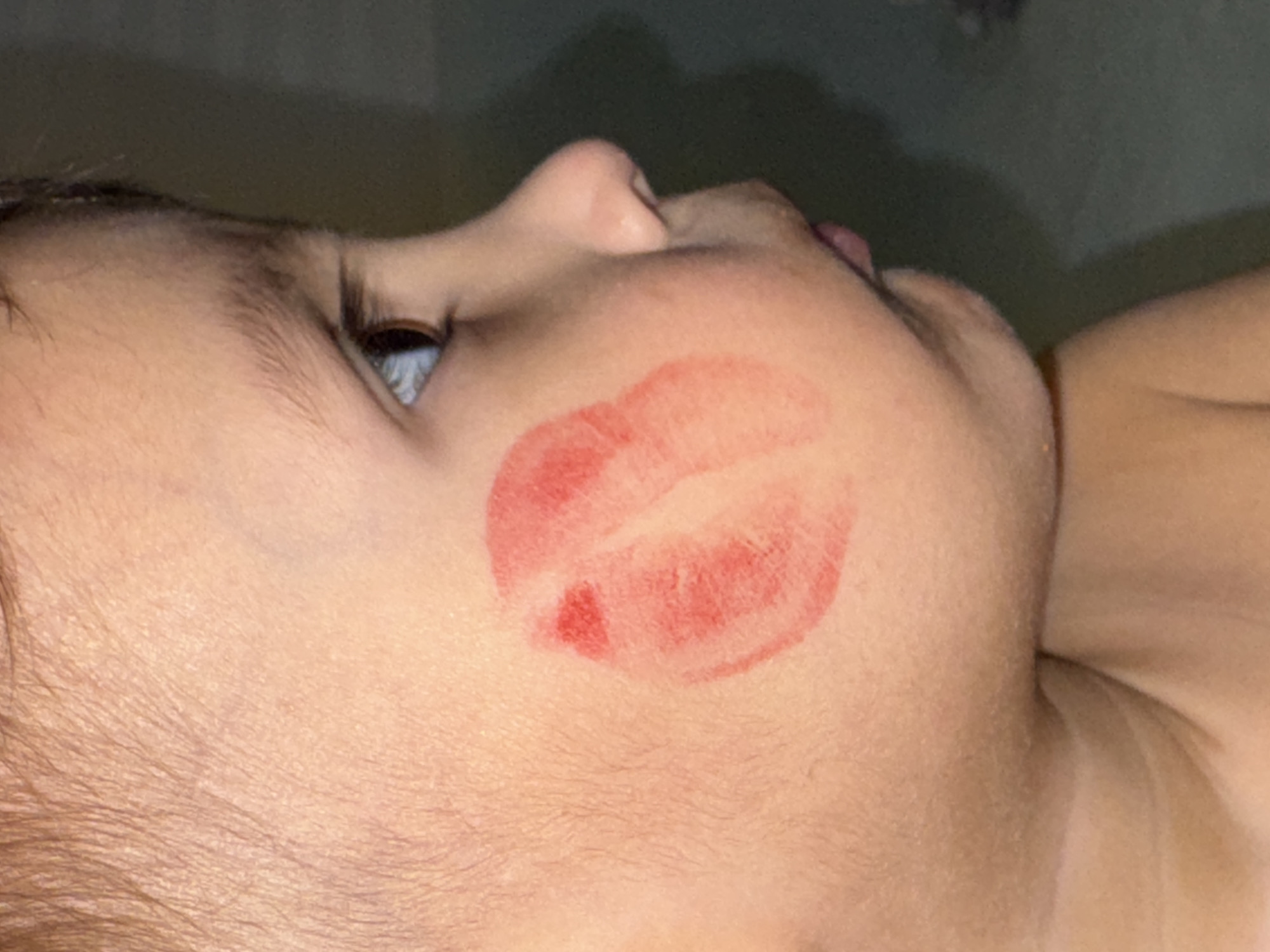 The image depicts a close-up of a baby's face with a red lipstick kiss on the cheek. The baby is lying on their side. The location is not identifiable from the image.