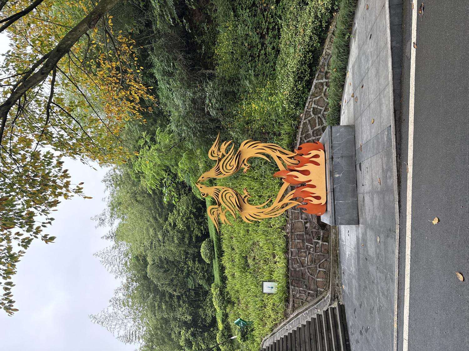 The image displays an outdoor scene, likely in a park or along a scenic pathway, characterized by lush vegetation and a prominent decorative sculpture. In the mid-ground, a golden-yellow sculpture features two stylized phoenix-like birds facing each other, their bodies integrated with vivid orange flame designs. This sculpture rests upon a dark grey, rectangular pedestal.
To the right of the sculpture, a grey paved sidewalk leads to an asphalt road, delineated by a white line. Scattered dry leaves on the ground suggest an autumn season. To the left, a stone retaining wall supports a steep, grassy incline densely covered with various green foliage, including trees and what appears to be bamboo. A large tree with distinct yellowing leaves frames the upper-left corner of the image, reinforcing the autumnal theme. A set of steps is partially visible descending along the retaining wall in the lower left. A small, green sign is present on the grassy slope, but any text on it is illegible.
The sky is bright and overcast, indicating daytime conditions with soft, diffused light. No people are visible, and no specific activity is taking place; the scene is static. The artistic style of the sculpture and the dense greenery are consistent with the provided location context of Liangjiang, China.