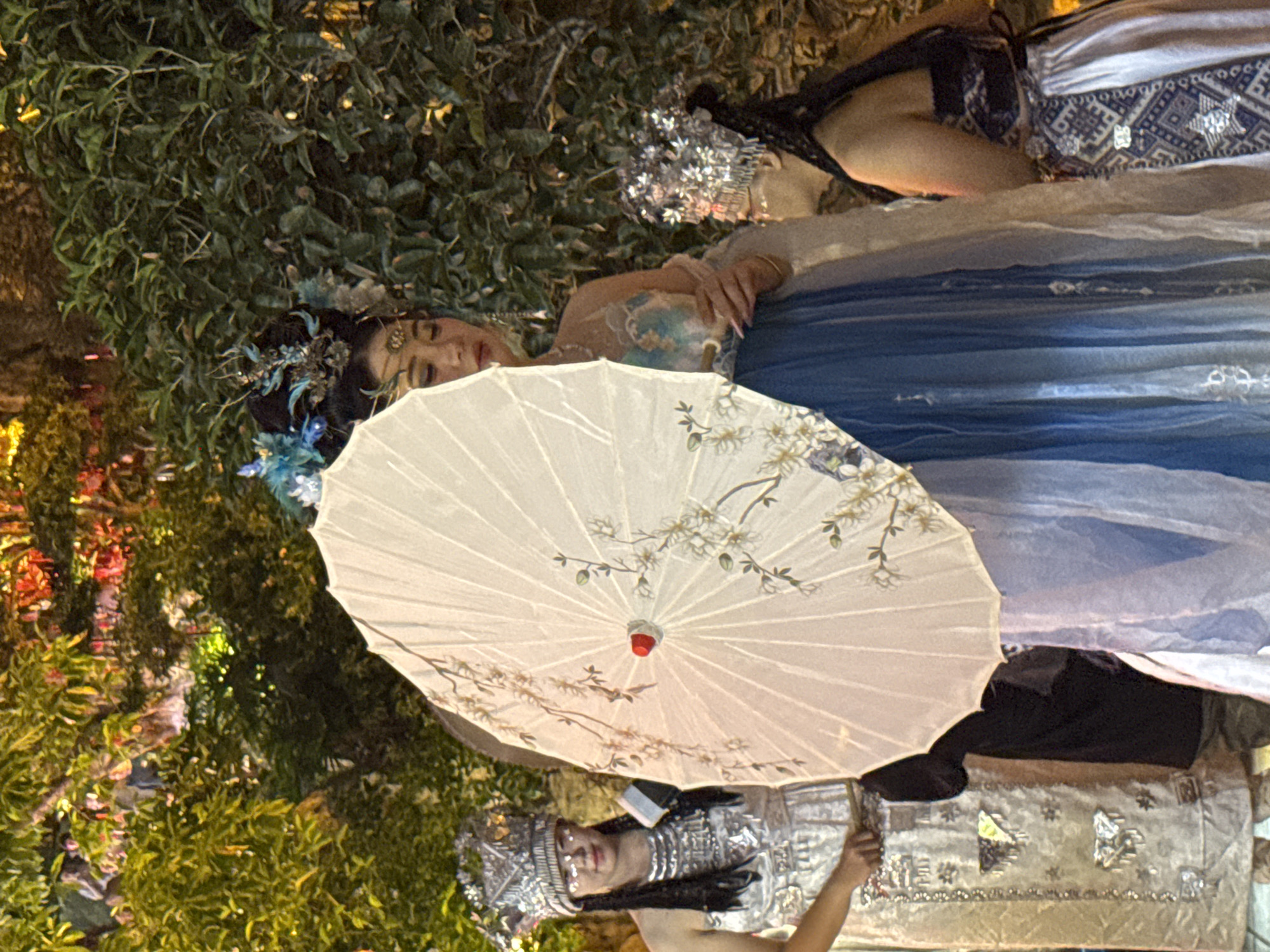 In the city of Yuzhong District, China, three women are posing for a photograph. The woman in the center is holding a large, white, paper umbrella decorated with floral motifs. She is wearing a blue and white dress with intricate embroidery and a blue and gold headpiece. To her left, a woman in a silver, ornate headdress and a sparkling, silver, sleeveless dress is standing. To her right, another woman in a silver, ornate headdress and a blue and white patterned dress is leaning in towards the center woman. The background is a lush green foliage wall, illuminated by warm, ambient lighting that suggests an evening setting, possibly a garden or outdoor event.