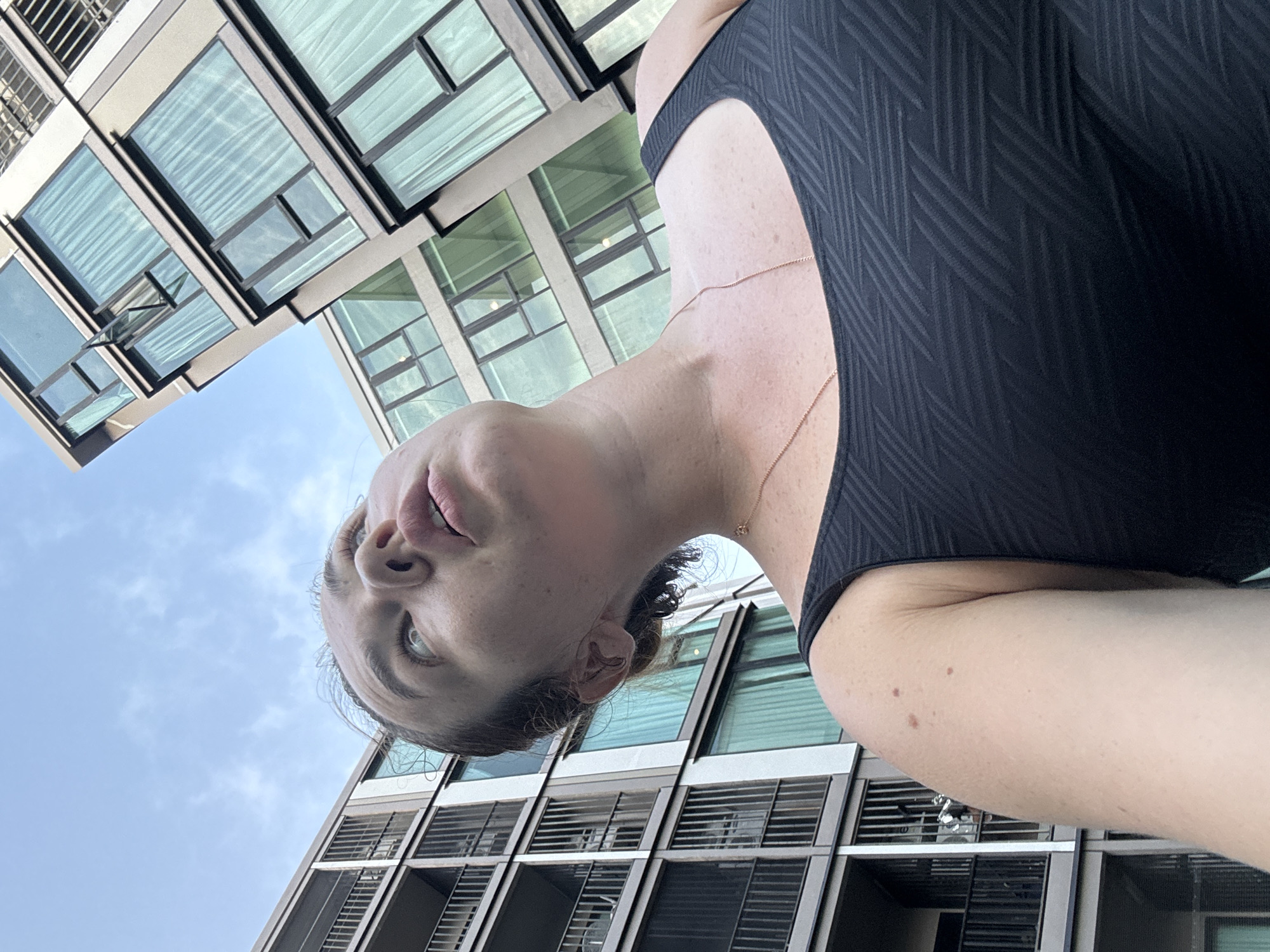 A woman is pictured from a low angle looking up at a modern building with many windows in Ratsada, Thailand. She is wearing a black tank top and a thin gold necklace. The building is tall and has multiple balconies with railings. The sky is cloudy and a light blue.