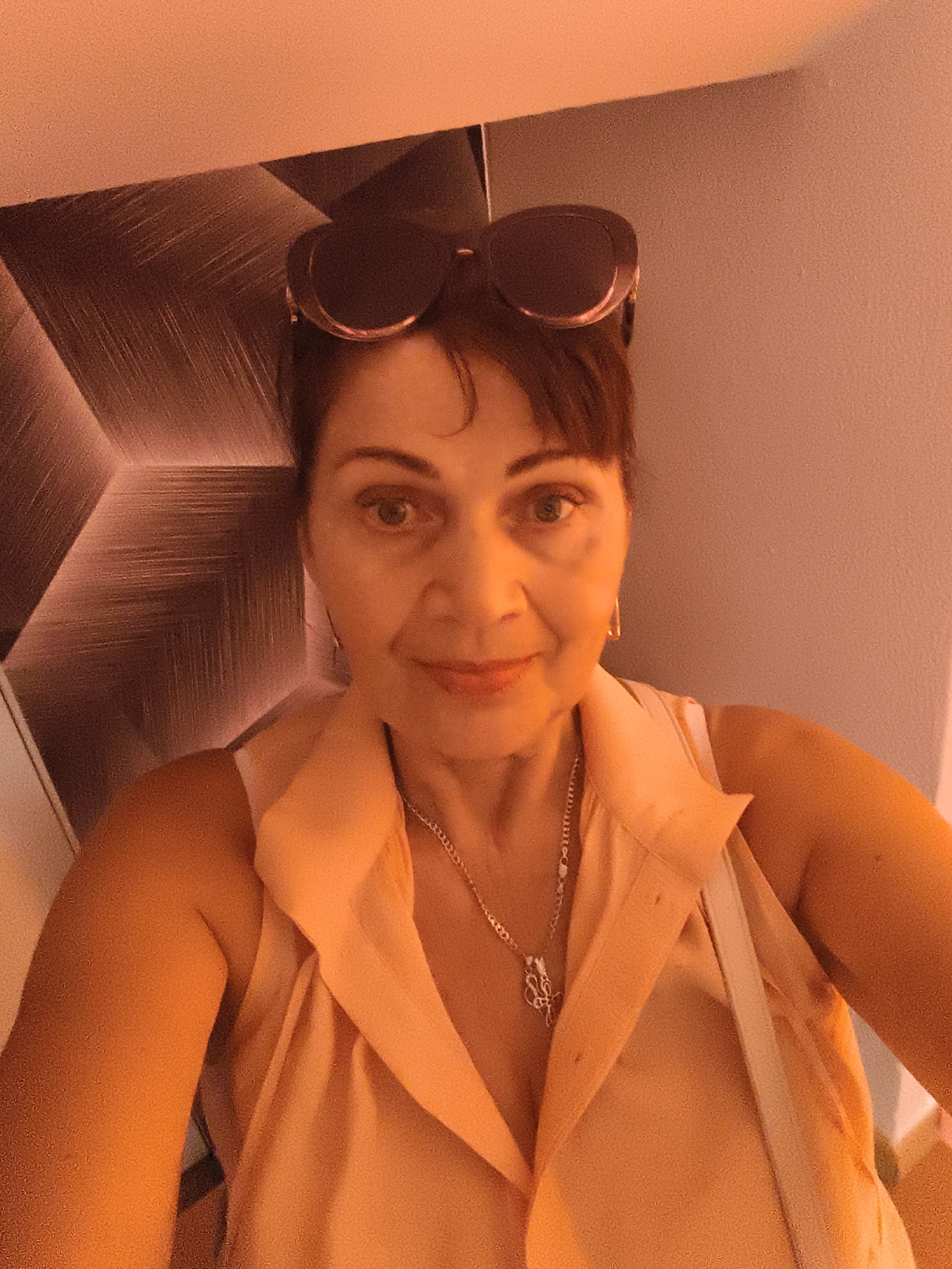 This is a selfie of a woman with dark hair, wearing sunglasses on her head and a light-colored sleeveless shirt. She is also wearing a silver necklace. The background is a wall with a geometric pattern and a plain wall. The lighting suggests it might be indoors, possibly in the evening or under artificial light, as the overall tone of the image is warm and orange-hued. The woman has a neutral expression. There is no visible text. Given the location context of Livorno, Italy, this could be a casual photo taken during a visit or while out and about.