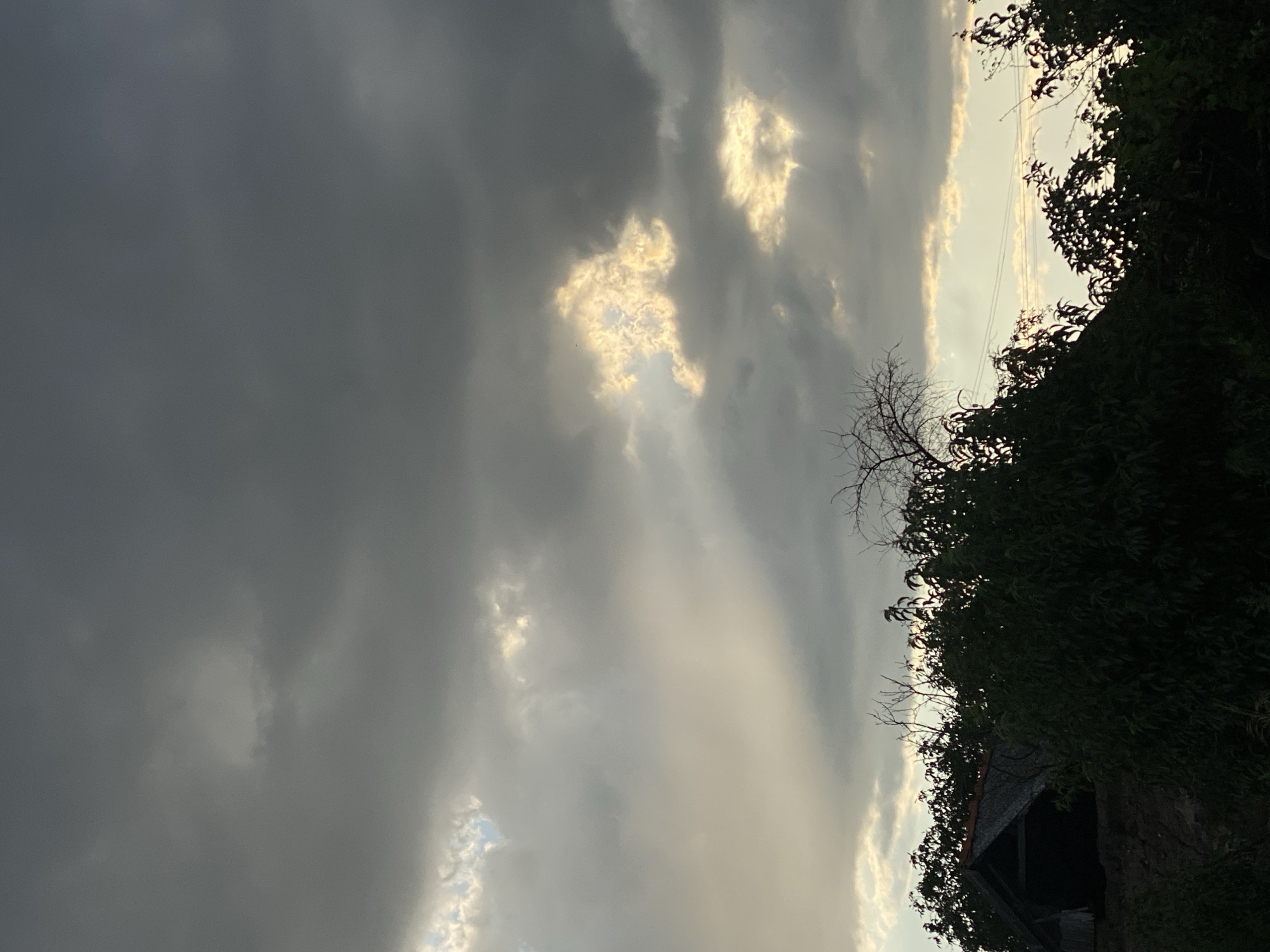 The image depicts a dusky sky over Cairaclia, Moldova, with dramatic, dark clouds partially illuminated by shafts of sunlight. In the foreground, silhouetted trees and dense foliage obscure a rustic, dark-colored structure, possibly a small shed or dwelling, with a weathered roof. The overall impression is one of an impending storm or the aftermath of one, with the natural elements dominating the scene. There are no people or animals visible.