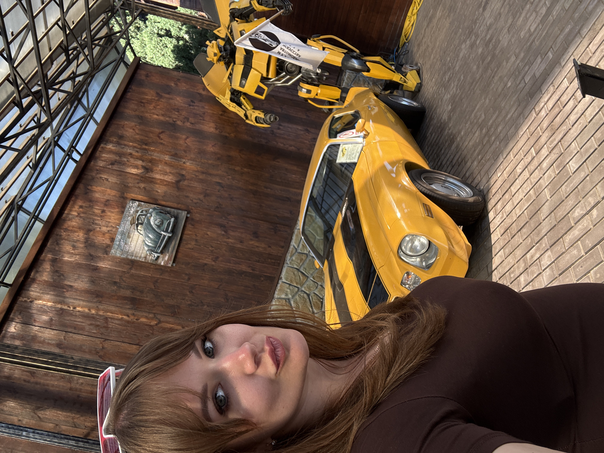 The image is a selfie of a woman with brown hair and light eyes, wearing a brown outfit. She is positioned in the foreground with a slight tilt to the left. Behind her is a yellow sports car and a yellow transformer sculpture, both suggestive of the Transformers character Bumblebee. The background also includes a wooden wall with a piece of artwork and a brick wall. A metal framework can be seen above, possibly part of a building's structure. The location is identified as город Ялта, Ukraine.