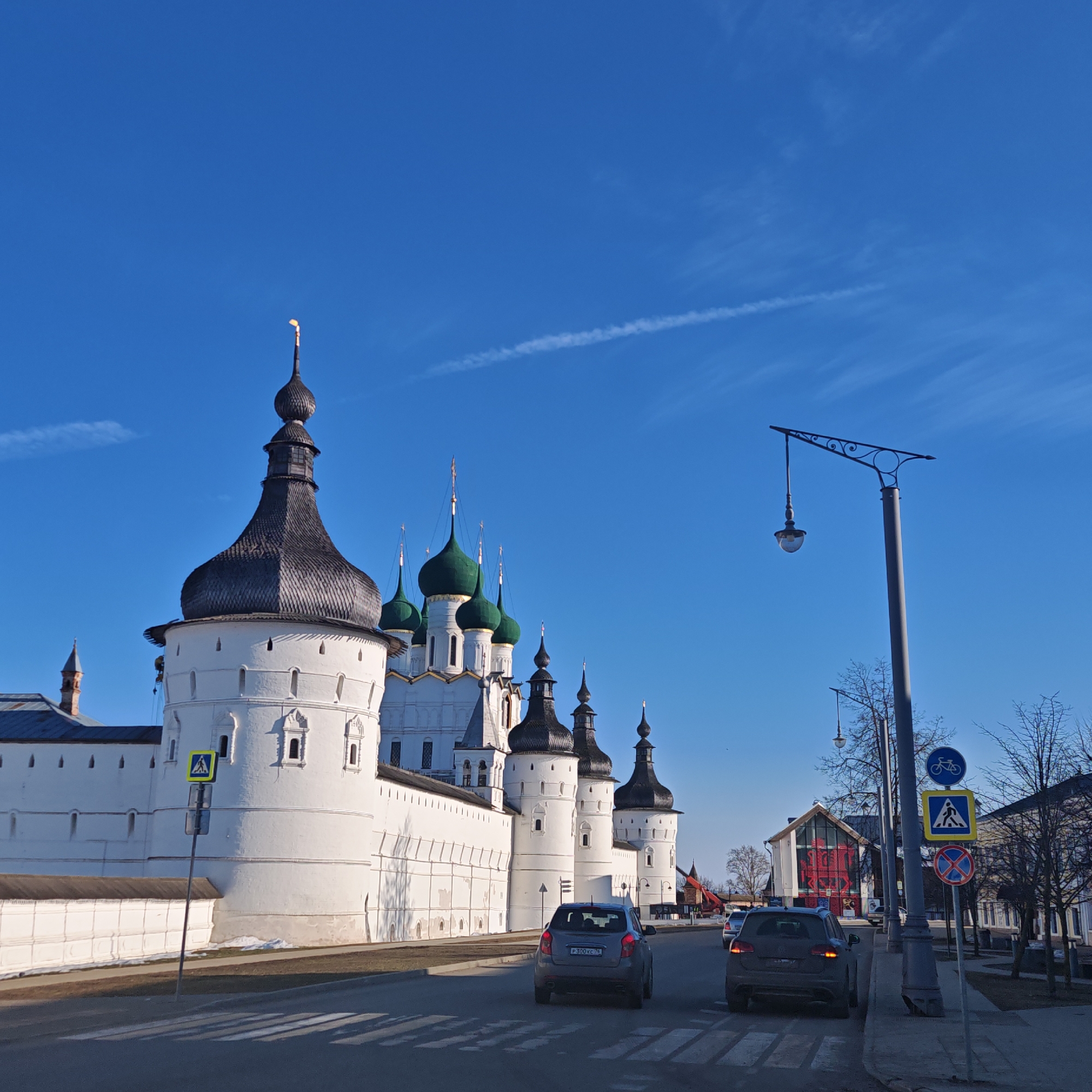This image depicts a street scene in Rostov Veliky, Russia, featuring a prominent view of the Rostov Kremlin complex under a clear, deep blue sky with faint cirrus clouds and a contrail.
In the foreground, an asphalt road with white zebra crosswalk markings is visible. Two vehicles are present: a dark grey hatchback, license plate P300KC 76, is positioned in the left lane, and a darker grey SUV is further back in the right lane. Both vehicles appear stationary or in slow transit.
To the left, the massive white-plastered outer wall of the Rostov Kremlin dominates the midground. The wall is fortified with several circular towers, each topped with a dark, conical, or onion-shaped roof; the most prominent tower on the far left features a golden finial. Behind the wall, several white-walled church structures with multiple green onion domes, crowned with golden spires and Orthodox crosses, are discernible. Patches of brown grass and remnants of snow are visible along the base of the Kremlin wall. A yellow square pedestrian crossing sign on a pole is positioned near the wall's base.
To the right of the road, a tall, dark grey street lamp pole with an ornate, curved arm holding a single downward-facing lantern stands. Below it, a vertical pole displays three road signs: a circular blue sign with a white bicycle symbol, a square yellow sign with a black pedestrian symbol, and a circular red 'no parking/stopping' sign. Behind these, several bare deciduous trees line a sidewalk, and further back, various urban buildings are visible, including one with a distinctive red façade or artwork behind a large window-like opening. No individuals are visible in the scene.