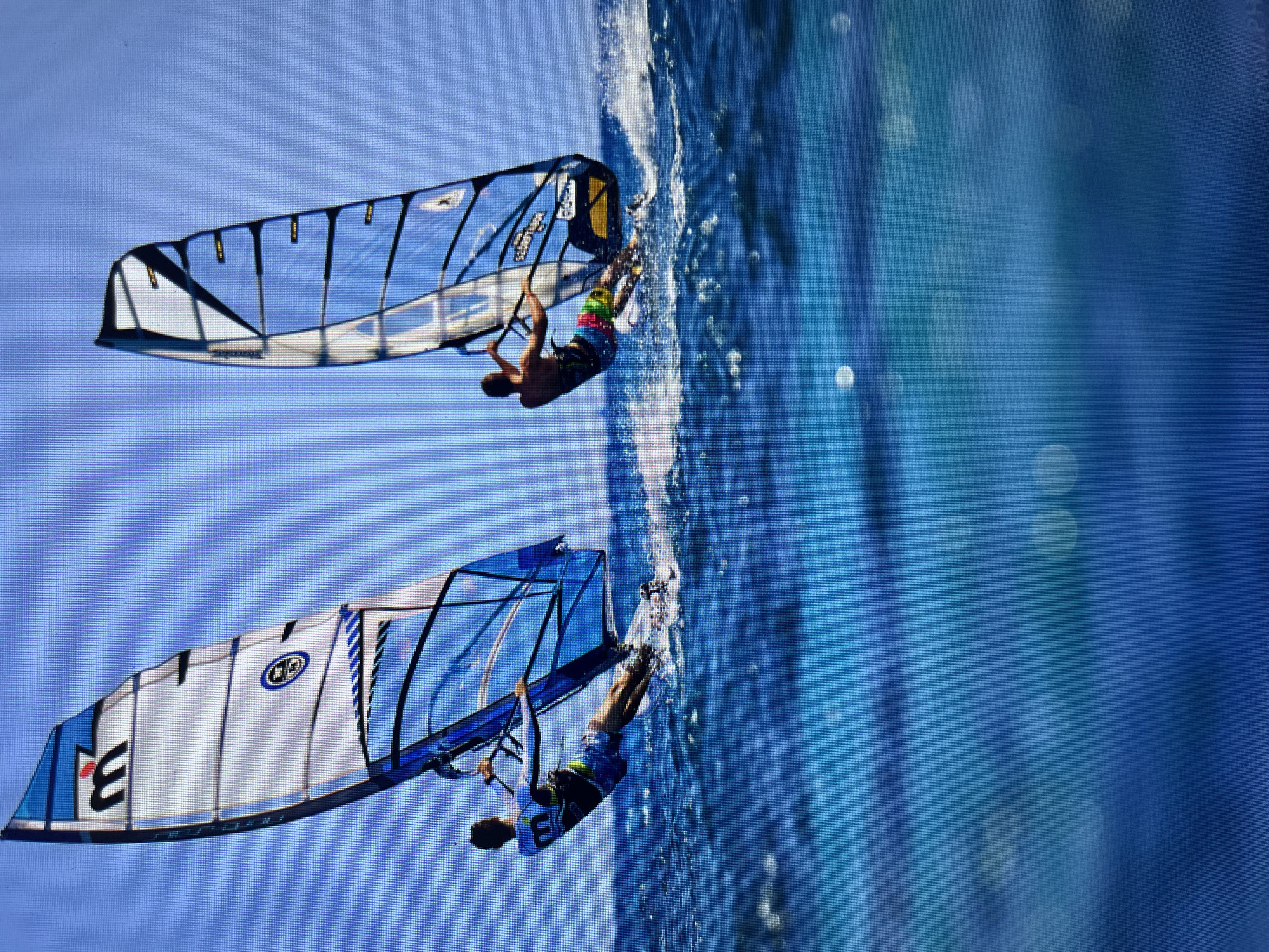 The image shows two windsurfers performing tricks. The water is churning behind them, indicating speed. The background is a clear, blue sky. The upper windsurfer wears colorful shorts, while the lower one sports a white shirt with a "3" on it and dark shorts. The sails of the windsurfers are blue and white. The upper sail has a yellow and black section, while the lower sail has black stripes. The location is Bacong, Philippines.
