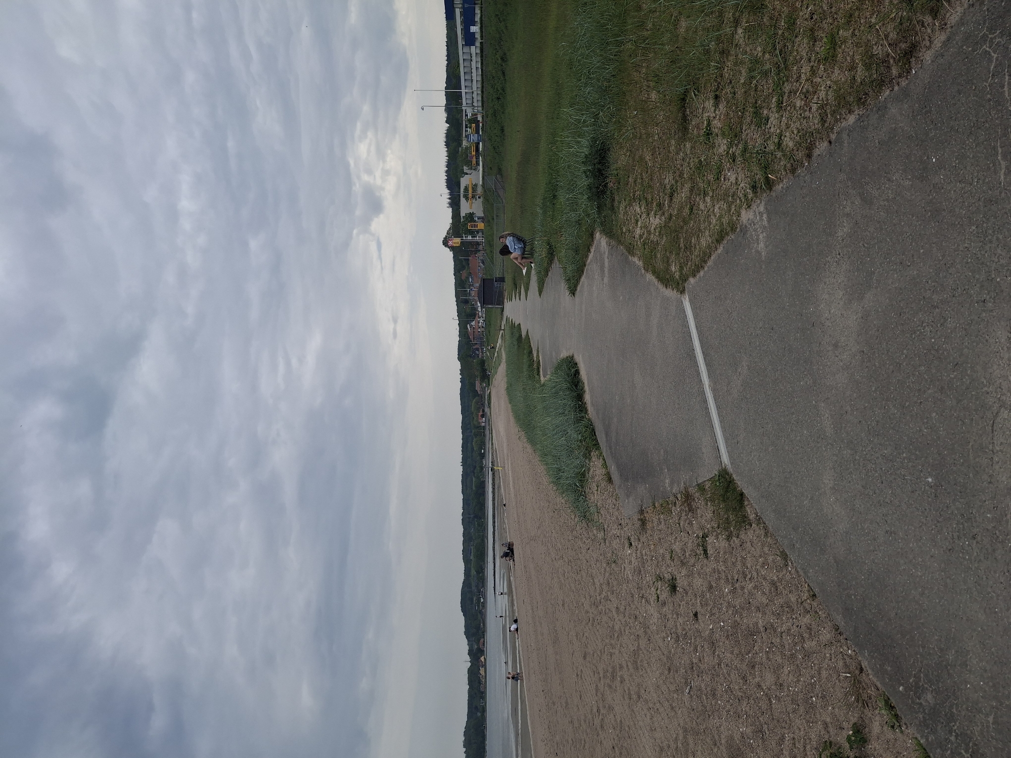 The image shows a sideways view of the beach at Aabenraa, Denmark. The sky is overcast with thick grey clouds. The shoreline extends along the left side of the image. On the right, a paved path runs parallel to the beach, separating it from a grassy area. People can be seen on the beach, some near the water's edge. Further down the path, two people are sitting on a bench. In the distance, there are buildings and trees, indicating the edge of the town.