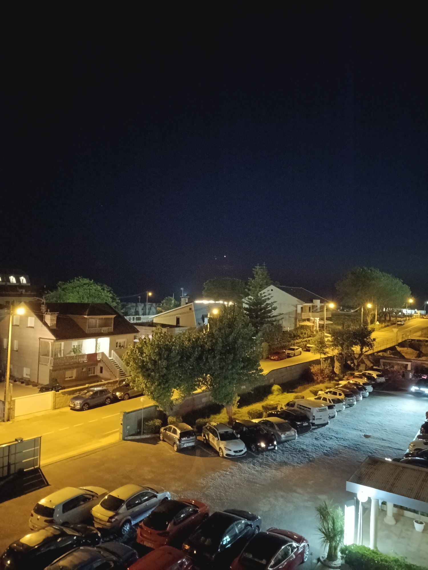 The image depicts a nighttime view of a residential area with parked cars and buildings. The scene appears to be from an elevated perspective, possibly a balcony or window, looking down onto a street and a parking lot.
In the foreground, a parking lot is filled with various cars, mostly sedans and some SUVs, illuminated by streetlights. The cars are of different colors including black, silver, white, and red. Some of the cars are parked in designated spots, while others are more loosely arranged.
Behind the parking lot, a street runs horizontally, also lined with parked cars. The street is illuminated by yellow streetlights, casting long shadows.
Several buildings are visible. On the left side of the image, a multi-story residential building with a sloped roof and balconies is prominently featured. It is lit from within, suggesting occupancy. To the right of this building, there are other houses and structures, some with lit windows. Trees are scattered throughout the scene, with dense foliage, suggesting a somewhat green environment despite the urban setting.
The sky is dark, indicating nighttime, with a deep blue hue. A few faint stars or distant lights are visible in the upper part of the sky.
The overall impression is a quiet, residential neighborhood at night, with the artificial lights of streetlamps and buildings providing illumination.
The location is identified as A Lanzada, Spain.