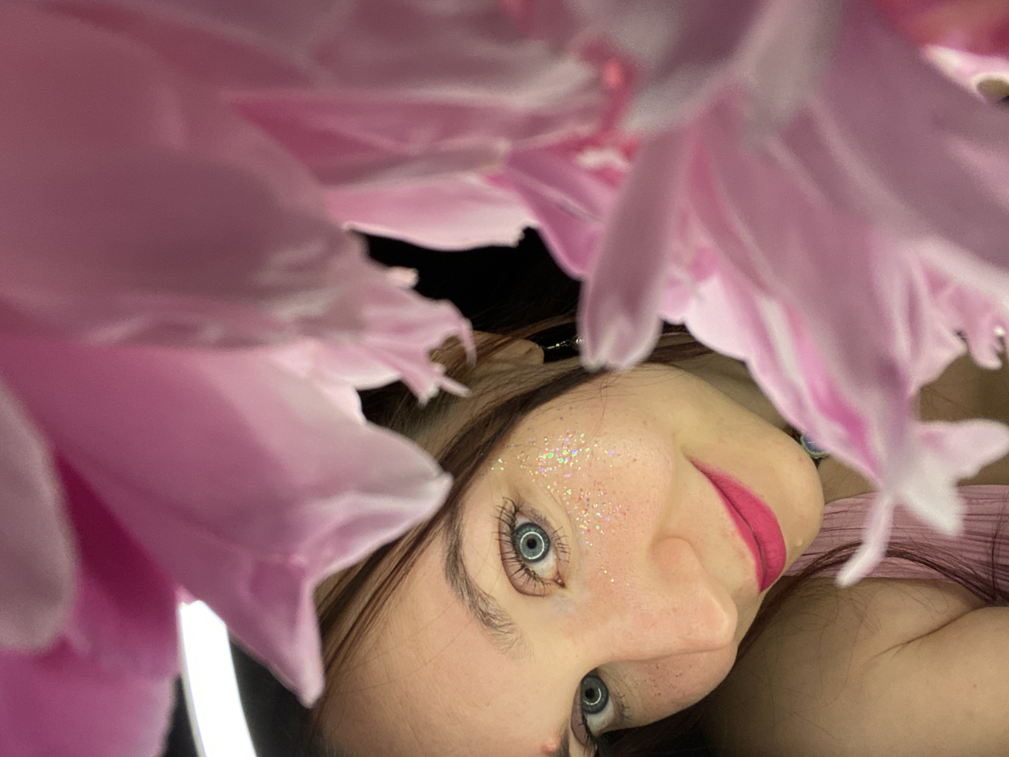 This media file presents a close-up, artistic portrait of a young woman, likely taken in Chaikovsky, Russia. The primary subject is the woman's face, partially obscured and intimately framed by large, soft pink flower petals, possibly from a peony. Her head is tilted, with her gaze directed upwards and slightly to the right, towards the viewer.
Notable details include her striking blue eyes, emphasized by dark eyelashes. Glitter is artistically applied to her forehead and cheekbone, catching the light. Her lips are painted a vibrant fuchsia pink, with a slight, gentle smile. Her dark hair frames her head. The petals in the foreground are soft and out of focus, enhancing the dreamy quality of the image. Reflections in her eyes and a visible vertical light strip in one of the crops suggest the use of artificial lighting, possibly a ring light, indicating a planned photoshoot. The background, where not covered by petals or hair, is dark. No discernible activity beyond posing for a portrait is taking place. There is no visible text. The overall mood is ethereal and beautiful, focusing on the woman's features and the soft floral elements.