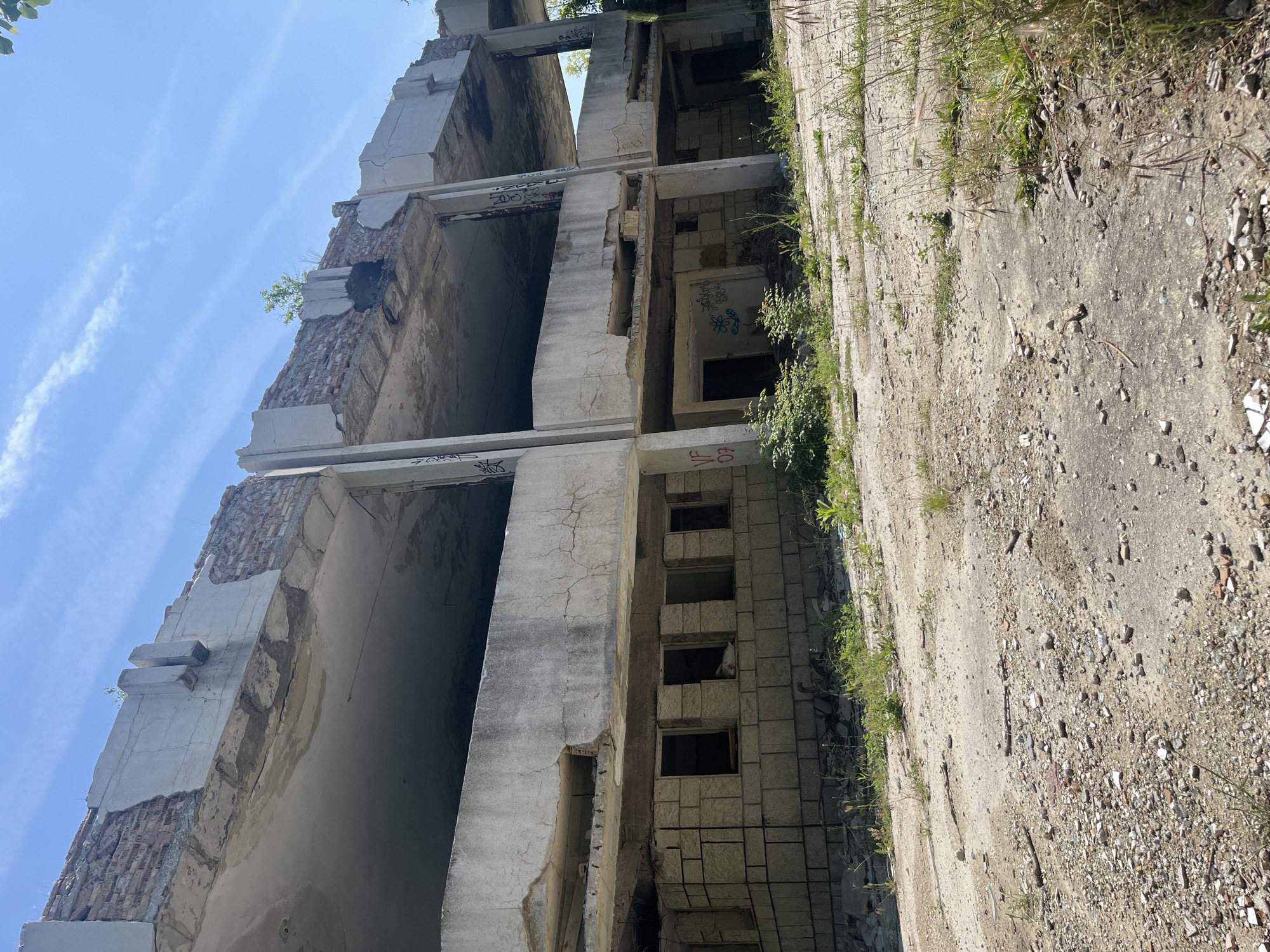 The image shows a multi-story, partially demolished concrete building in Varna, Bulgaria. The building lacks external walls, revealing its internal structure and partially collapsed floors. There's exposed brickwork in some sections. Graffiti is visible on some of the concrete beams and walls. The ground around the building is covered in rubble, dirt, and vegetation. The sky is blue with streaks of clouds. The picture is taken from a low angle, looking up at the building.