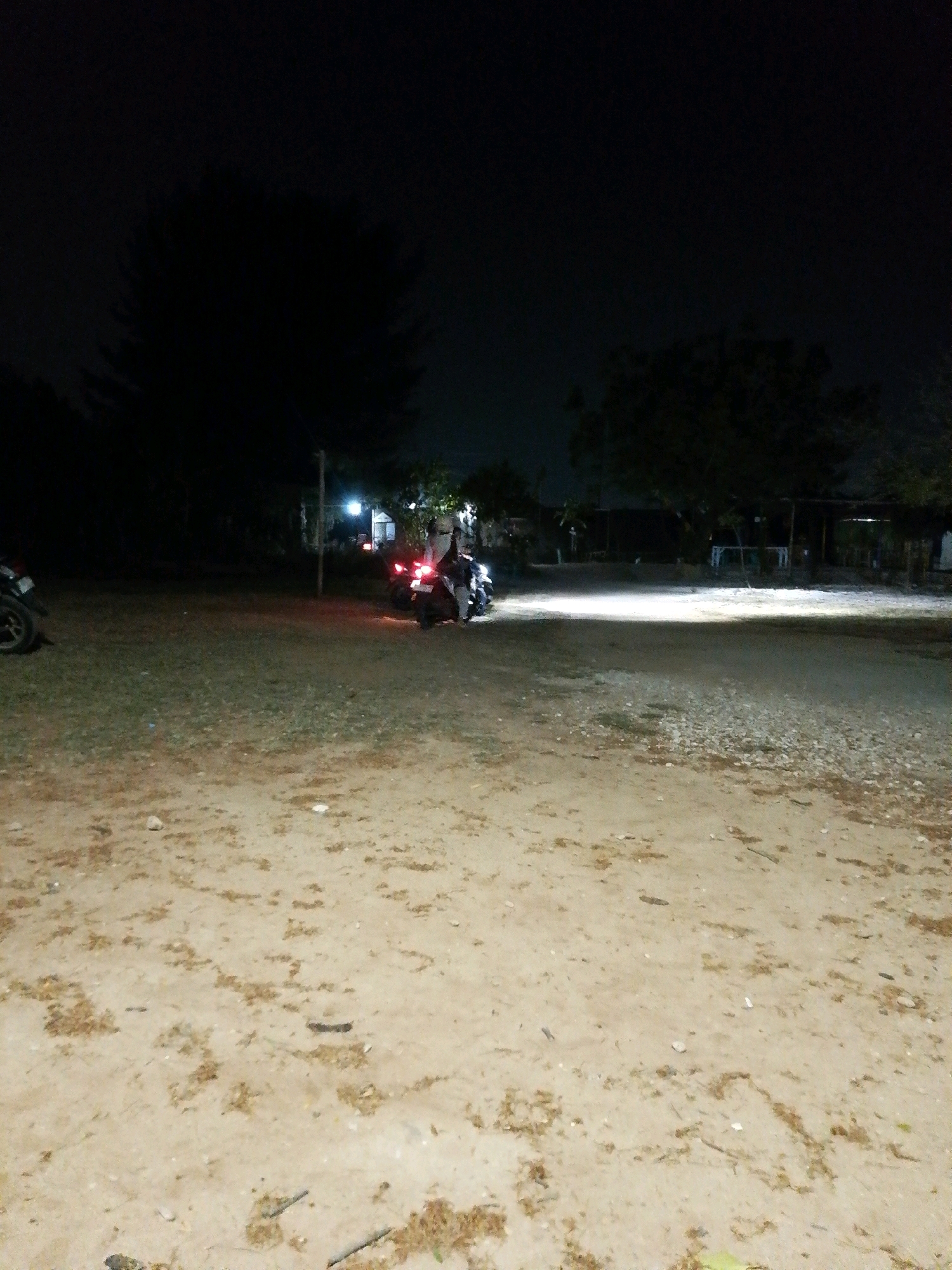 The image depicts an outdoor scene at night, likely in or around Cikal, Indonesia. The foreground is a dirt and gravel ground with some scattered dry leaves and debris. In the mid-ground, a dirt road or open area is illuminated by a bright light source, possibly from a building or a vehicle. To the right of this illuminated area, there are several structures that appear to be simple buildings or open shelters with visible lights. A motorcycle is parked on the left side of the frame, facing away from the camera. In the right mid-ground, two motorcycles are visible. One motorcycle has its brake lights illuminated. A person or people are near the motorcycles on the right, though their features are indistinct due to the low light conditions and distance. Trees and dark silhouettes of foliage are visible in the background, suggesting a rural or semi-rural setting. The overall atmosphere is dark, with artificial lighting creating distinct areas of brightness.