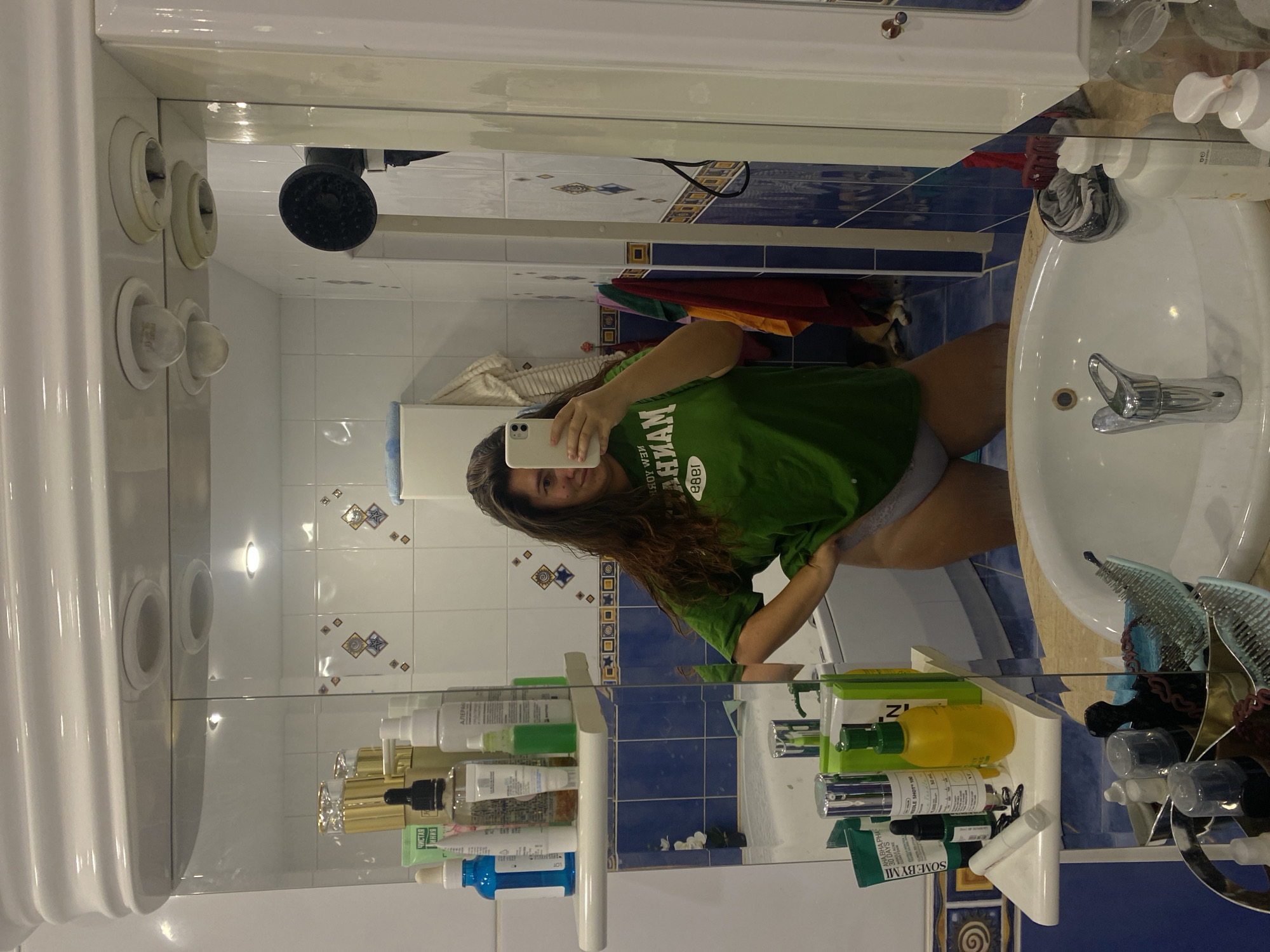 In a bathroom with blue tiled walls and white fixtures, a young woman with long, dark, wavy hair takes a mirror selfie. She is wearing a green t-shirt and grey underwear. Various toiletries and cosmetic products are visible on a shelf above the sink and on the counter. The overall impression is one of a casual, personal moment captured at home. The image does not contain any information that would allow for determining the city of Perm, Russia.