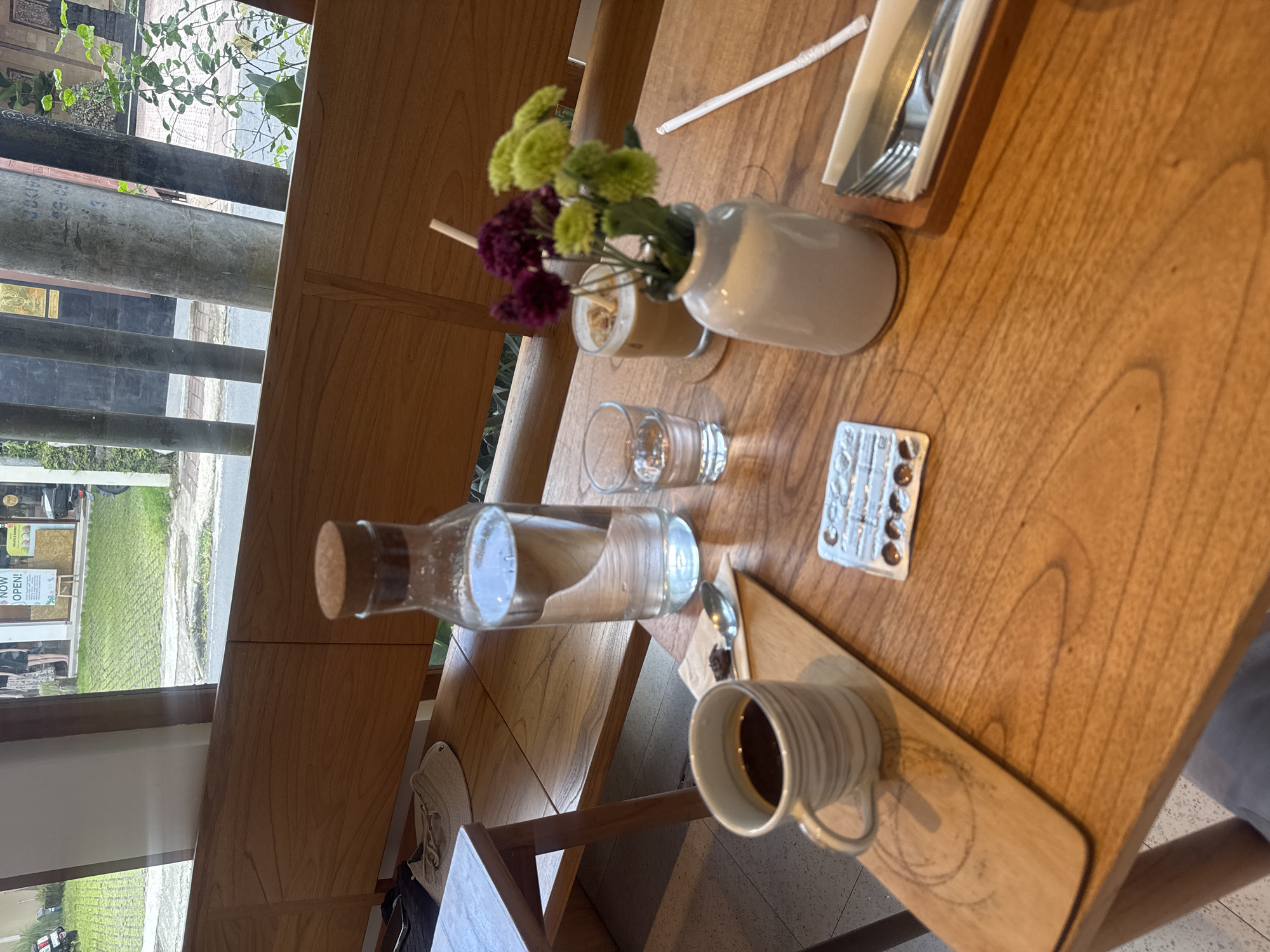 The scene shows a wooden table at a cafe or restaurant in Ubud District, Indonesia. Sunlight streams in through a window to the left, illuminating the table. Visible outside is vegetation, and the “NOW OPEN!” sign can be seen. On the table is a water bottle, a half-full glass of water, a small bouquet of flowers, cutlery in a holder, and a cup of coffee on a wooden tray. A blister pack of pills is present. A chair and a glimpse of a person's leg can be seen at the bottom right.