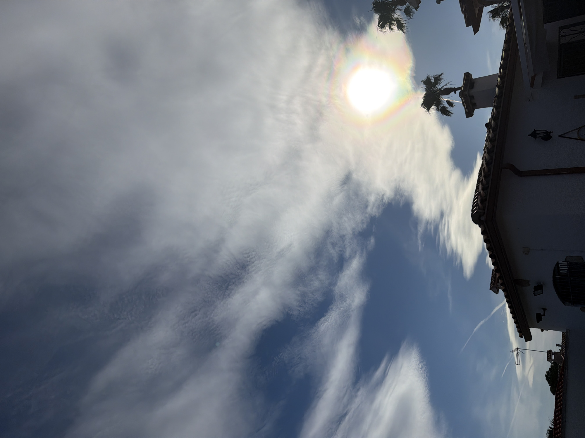The image displays a vibrant daytime sky over Mont-roig del Camp, Spain, featuring architectural elements and natural phenomena. The primary subject is the bright sun, which is surrounded by a prominent, colorful solar halo, indicating the presence of ice crystals in the atmosphere. The sky is a mix of deep blue patches and dynamic white clouds, ranging from thick and diffuse to wispy, streaky formations.
To the right side of the frame, a segment of a building with white stucco walls and a reddish-brown tiled roof is visible. Its architecture, including a decorative outdoor light fixture, a dark drainpipe, and a window with security bars, evokes a Mediterranean or Spanish style. Several dark silhouettes of palm trees are scattered across the upper portion of the frame, some appearing near the sun and others above the building. A distinct chimney with an ornate cap rises from the roofline. Below the main roofline, an antenna or aerial is visible on a lower section of the roof. A faint white contrail from an aircraft can be seen cutting across a patch of blue sky. The scene suggests a sunny, pleasant day. No text is visible in the image.
