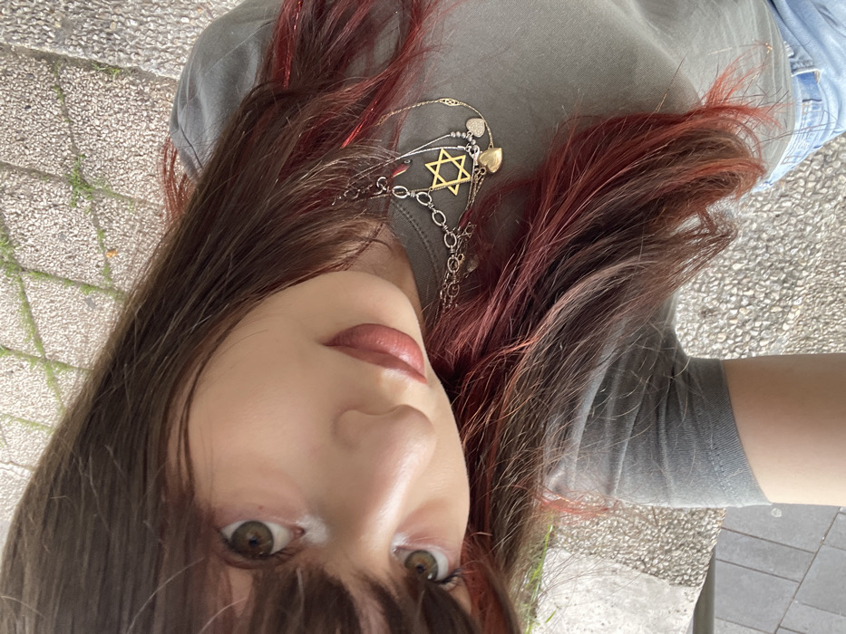 A close-up, high-angle portrait shows an individual with brown hair featuring reddish highlights and bangs, looking directly into the camera. The individual wears a grey short-sleeved t-shirt and partially visible blue jeans. Around the neck are multiple layered necklaces, prominently featuring a gold-colored Star of David pendant, a silver-colored chain, and at least two heart-shaped charms (one gold-colored, one silver-colored). The background consists of light-colored paving stones with visible green foliage in the cracks, indicating an outdoor setting in the city of Raanana, Israel.