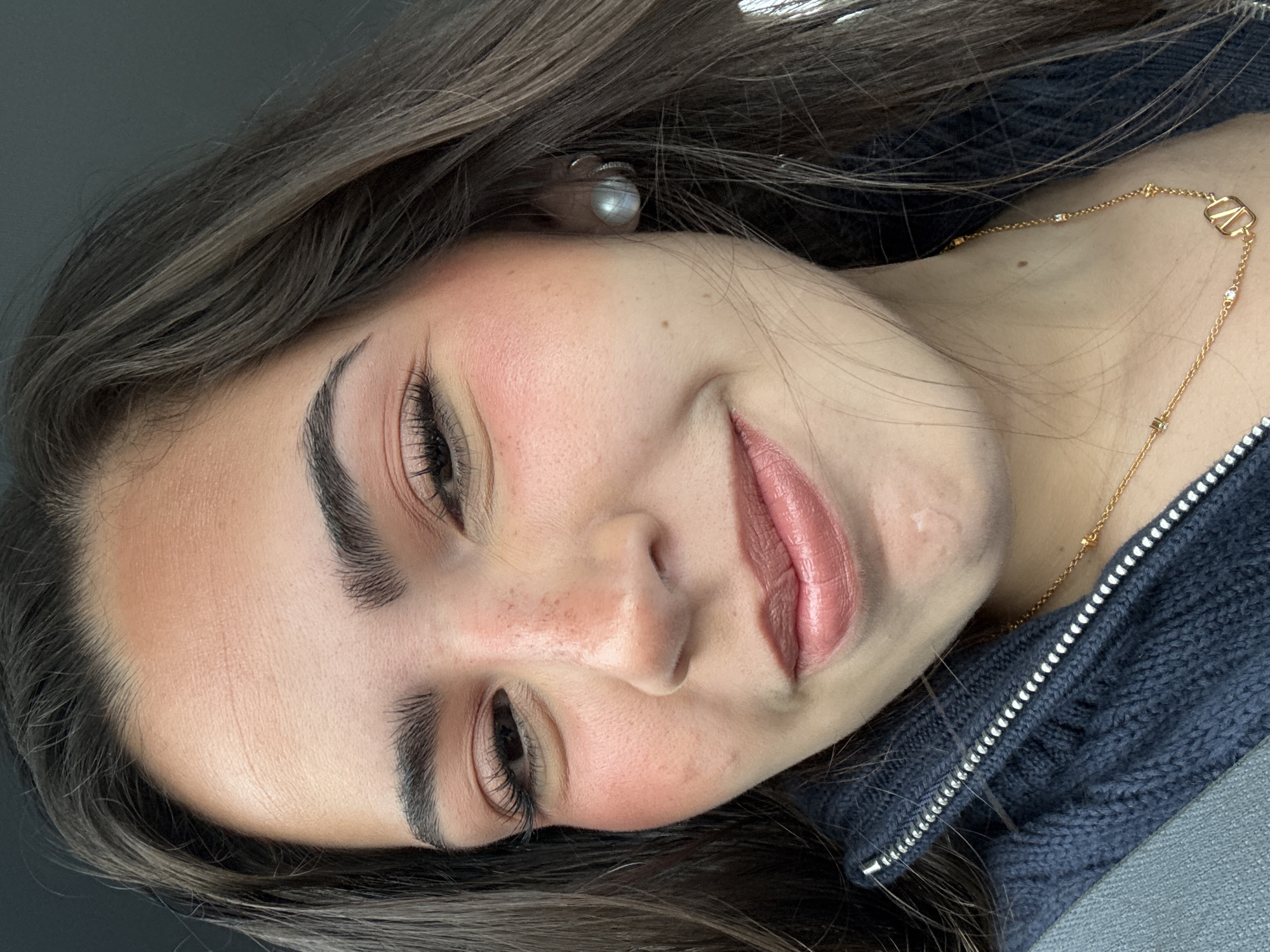 A medium shot depicts a young woman with fair skin, brunette hair, and professionally applied makeup, including defined eyebrows, eyeshadow, blush, and lipstick. She is wearing a pearl earring and a gold necklace. She is wearing a dark-colored zippered sweater. The location is presumed to be San Antonio, United States of America.
