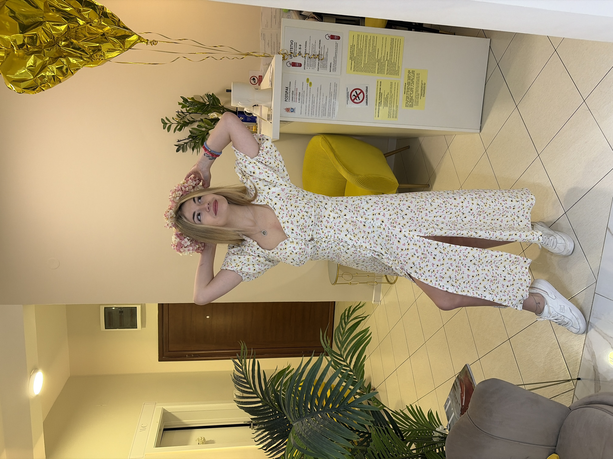 A woman in a floral dress and sneakers stands indoors, seemingly posing for a photo. She is wearing a flower crown. Behind her are decorative plants, a yellow chair, and a cabinet with warning signs written in a language other than English. A golden balloon is also visible. The location appears to be a public space with tiled floors.