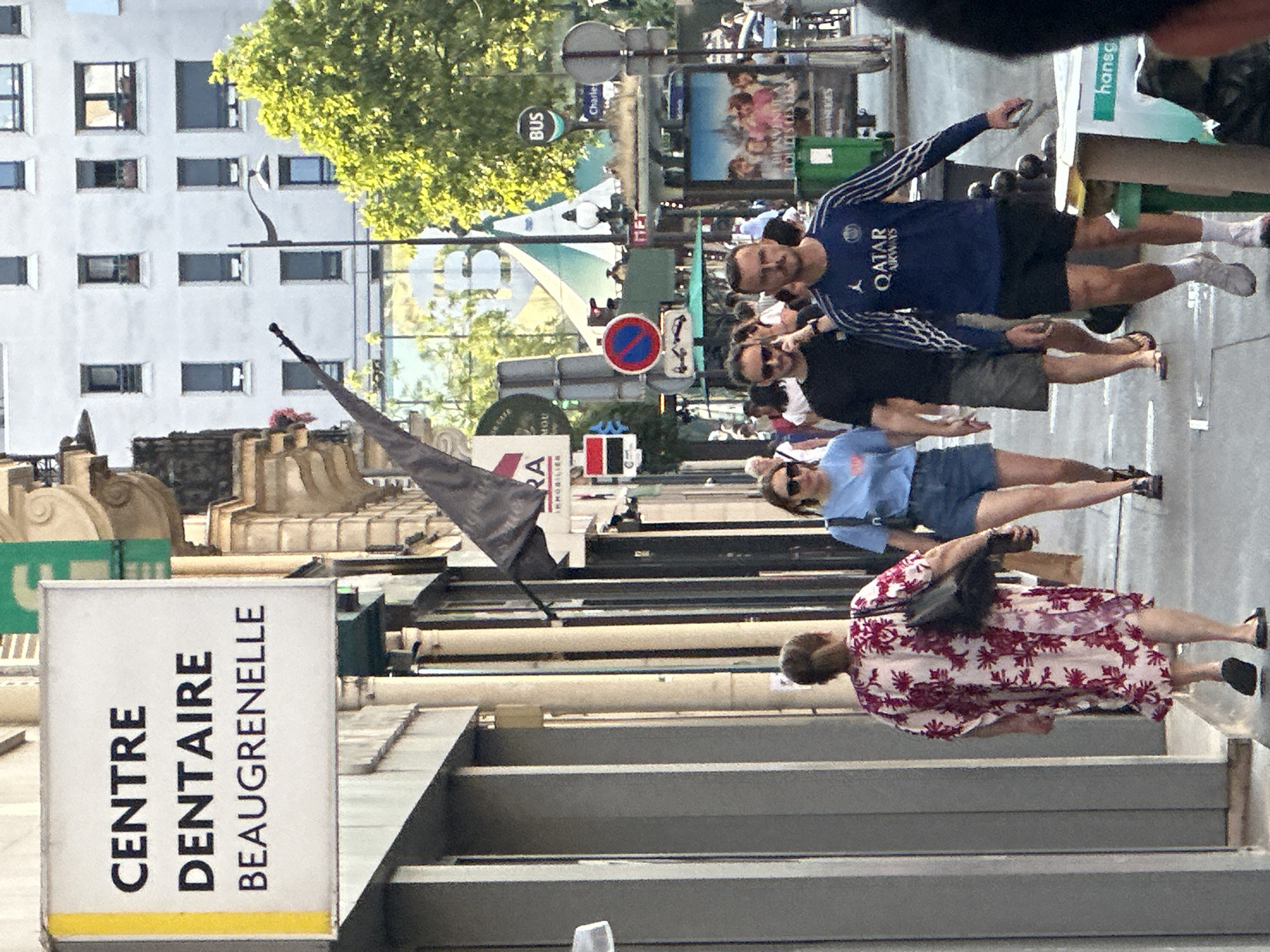 The image depicts a street scene in Paris, France, likely during daylight hours, given the lighting conditions. In the foreground, four individuals are walking on a sidewalk. A woman in a pink and white floral dress is walking away from the camera, carrying a dark bag. To her right, a woman wearing a blue t-shirt and denim shorts is walking towards the camera, also holding a bag. Further to the right, two men are walking towards the camera. The man on the left is wearing a black t-shirt and grey shorts, while the man on the right is wearing a blue long-sleeved shirt with white detailing and black shorts.
A sign with yellow trim is visible on the left side of the image, bearing the text "CENTRE DENTAIRE BEAU GRENELLE" in black capital letters. This indicates the presence of a dental center with that name. In the background, a building with multiple windows is visible, suggesting an urban environment. Several street signs are present, including a circular red and blue sign with a white bar and a white exclamation mark, indicating a traffic regulation. A "BUS" sign is also visible. Trees with green foliage line the street. A street vendor's cart or kiosk is partially visible on the right side of the image. The overall scene is that of a busy city street with pedestrians.