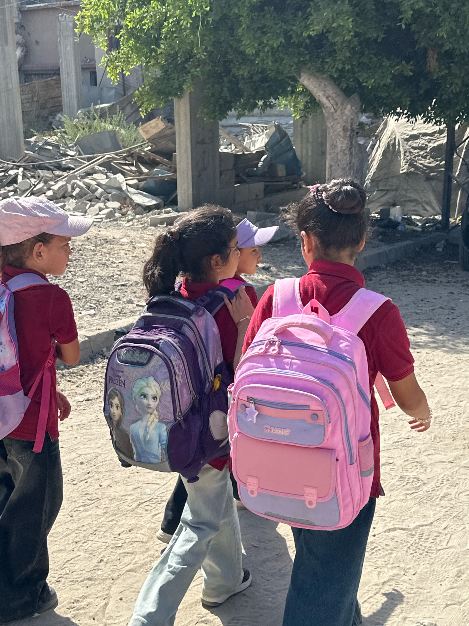 In Gaza City, Palestinian Territories, three young girls in school uniforms walk on a dirt road, carrying backpacks. The girl on the left, wearing a pink cap and a maroon shirt, has a pink backpack. The middle girl, also in a maroon shirt, carries a purple backpack featuring characters from Disney's "Frozen." The girl on the right, facing away from the camera, wears a maroon shirt and a pink backpack. The background shows damaged buildings, debris, and tents, indicating a context of destruction. The lighting suggests it is daytime.