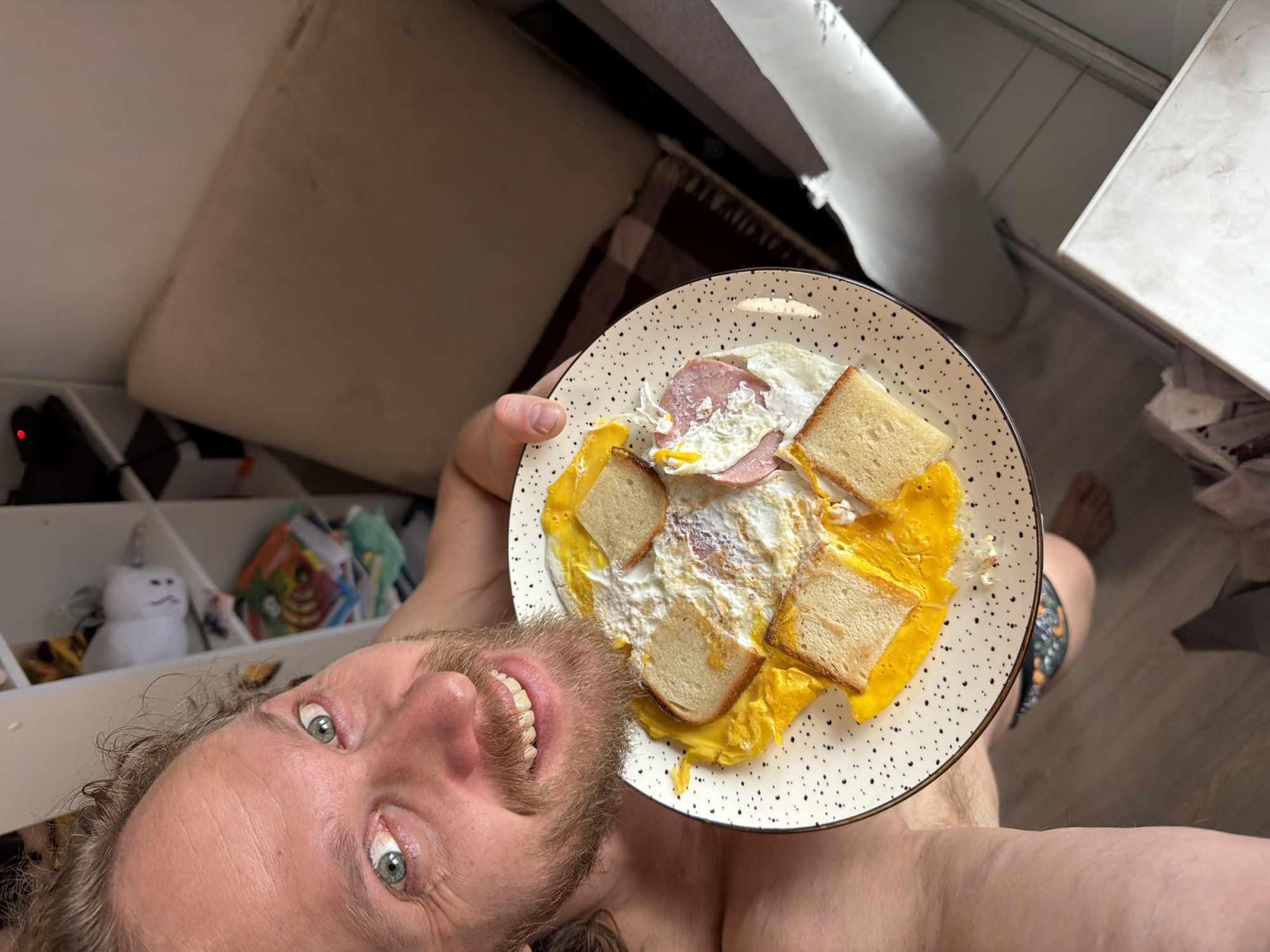 A man is holding a plate of eggs, toast, and ham, likely at home given the presence of furniture and personal belongings in the background. The plate is a speckled white with black dots. The man has light skin, light eyes, a beard and a visible smile. The background includes a white shelf with items such as stuffed animals and books, a sofa, and a section of tiled flooring. The location within Saint Petersburg, Russia, cannot be determined from the image.
