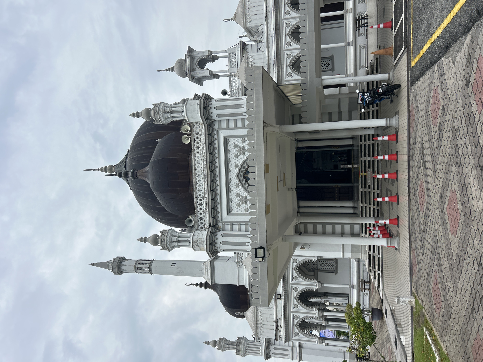 The image shows the exterior of Zahir Mosque in Alor Setar, Malaysia, on a cloudy day. The mosque is a grand structure with multiple domes and minarets, predominantly white with dark domes. In the foreground, a paved walkway leads to the entrance, bordered by red and white traffic cones. A motorcycle is parked on the right side of the walkway. A small tree in a pot is visible on the left side of the walkway. The architecture features intricate detailing, including arches and decorative patterns. The overall scene depicts a religious site with some temporary barriers and a parked vehicle.
