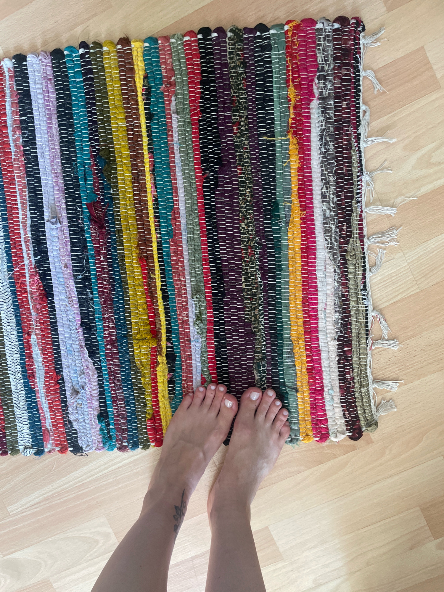 A pair of bare feet with white toenail polish is positioned on a colorful, braided rag rug placed on a light-colored wooden floor. The rug features vertical stripes of various colors including red, pink, purple, green, teal, yellow, brown, and black, with white stitching visible throughout. The feet are positioned near the bottom edge of the rug. The background is a simple wooden floor. There is a faint tattoo on the left shin. The location is specified as Atarfe, Spain.