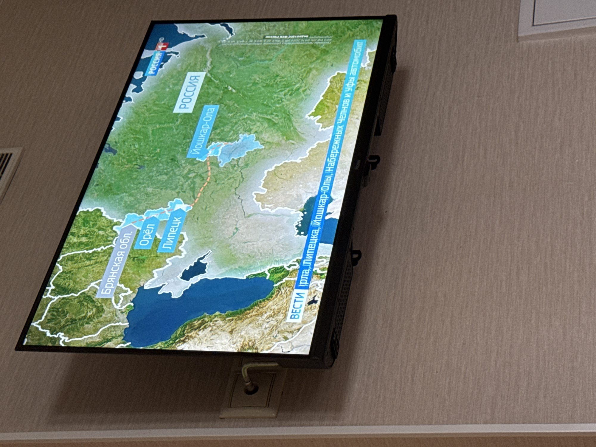 A widescreen television displays a map of Russia with several cities labeled, including Bryansk Oblast, Oryol, Lipetsk, and Yoshkar-Ola. Text overlays indicate the channel "Rossiya" with an "HD" logo. Another text overlay at the bottom of the screen reads "VESTI" followed by a partial sentence in Russian, which translates to "reports from Iran, Lipetsk, Yoshkar-Ola, Naberezhnye Chelny and Ufa by car." A power outlet and cable are visible to the left of the television, mounted on a beige textured wall. The TV is positioned at a slight upward angle. The image is taken from a slightly elevated perspective.