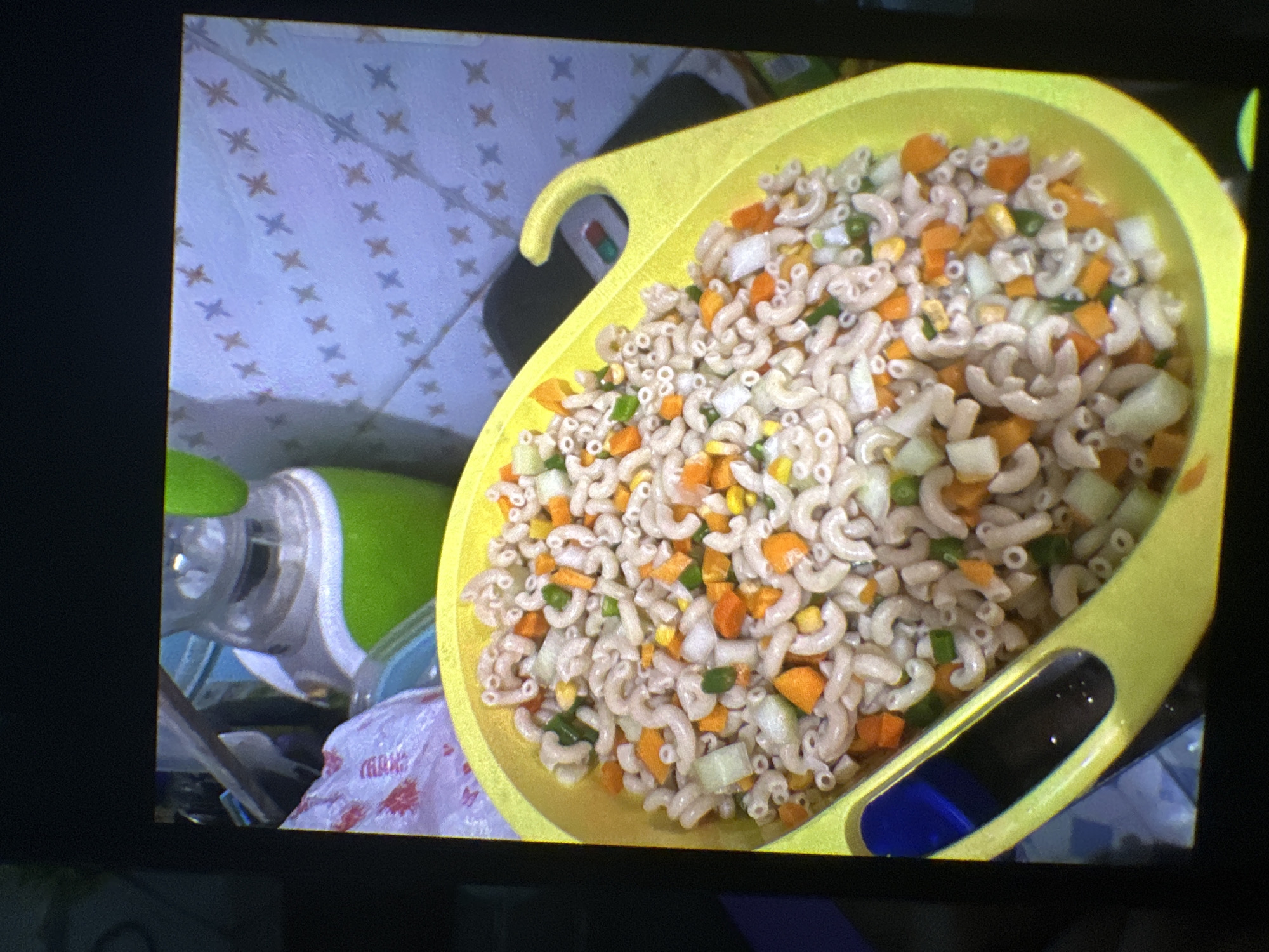 A bright yellow bowl is filled with macaroni salad mixed with diced carrots, celery, and corn. The bowl is resting on a dark surface, possibly a table, and is partially filled with water in its lower section, suggesting it might be a colander. In the background, a blender with a green top is visible, along with a patterned wall. The overall scene appears to be an indoor setting, possibly a kitchen, though specific details about the city of Rido, Nigeria, are not discernible from the image. There are no people or specific actions depicted in the image.