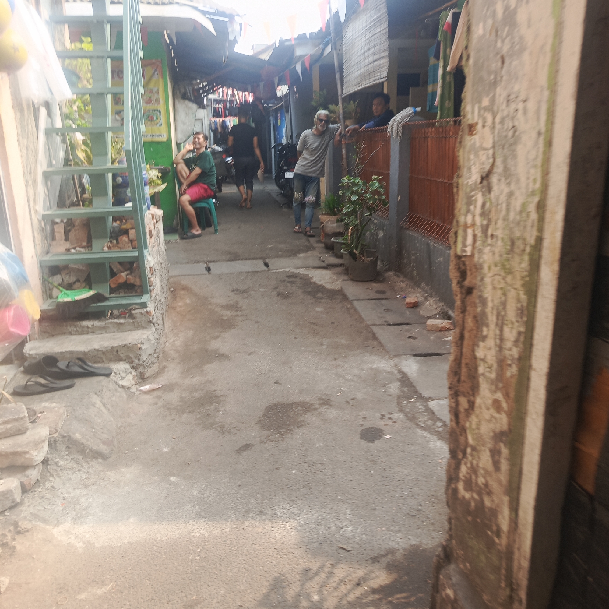 The image depicts a narrow alleyway in an urban environment, likely within the city of South Jakarta, Indonesia. The alley is lined with buildings and features a mix of residential and commercial elements.
In the foreground, a person is seated on a green plastic stool, wearing a green t-shirt and red shorts. They appear to be engaged in conversation or observing their surroundings. To the left, a set of outdoor stairs with a light blue railing leads up to a structure.
Further down the alley, another individual, an elderly man with white hair and sunglasses, leans against a fence. He is wearing a grey t-shirt and ripped blue jeans. Behind him, another person is visible, seemingly looking out into the alley.
The ground is paved with asphalt and shows signs of wear and tear, with some debris and puddles present. Various objects are scattered throughout the scene, including potted plants, a motorcycle, and what appear to be laundry items hanging from lines. The overall impression is one of a densely populated and active urban community.