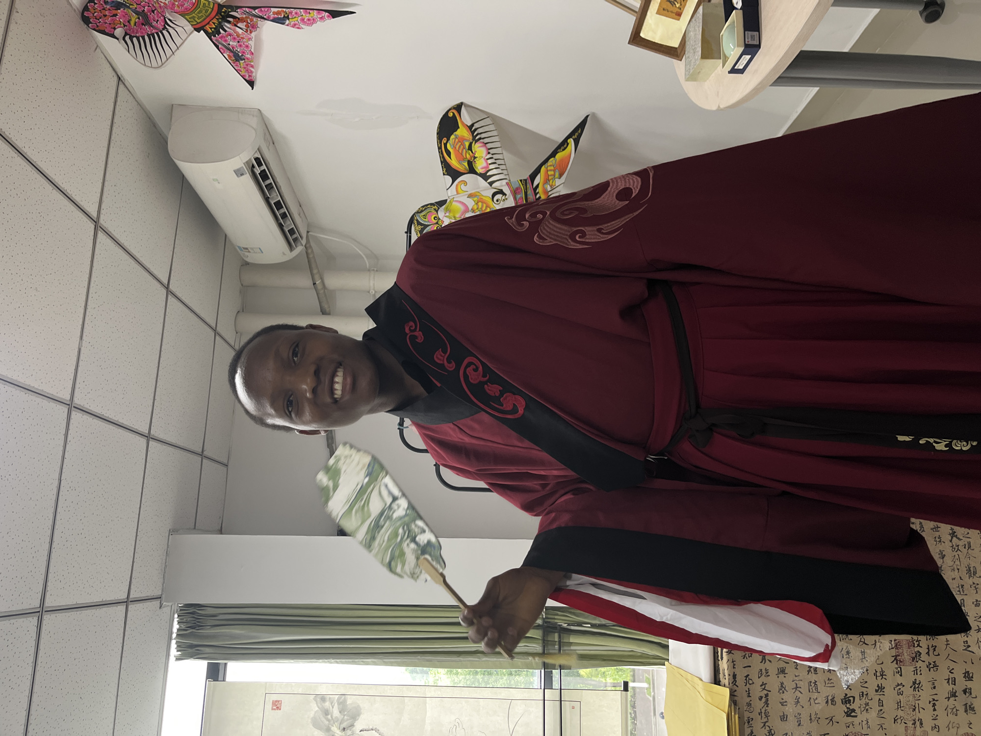 A man stands in a room in Jiangjin, China. He is wearing a maroon and black robe and holding a fan with a green marbled design. He is smiling and looking directly at the camera. Behind him, there is a window with green curtains, an air conditioning unit, decorative kites hanging from the ceiling, and a table with objects on it. Chinese calligraphy is visible in the background.