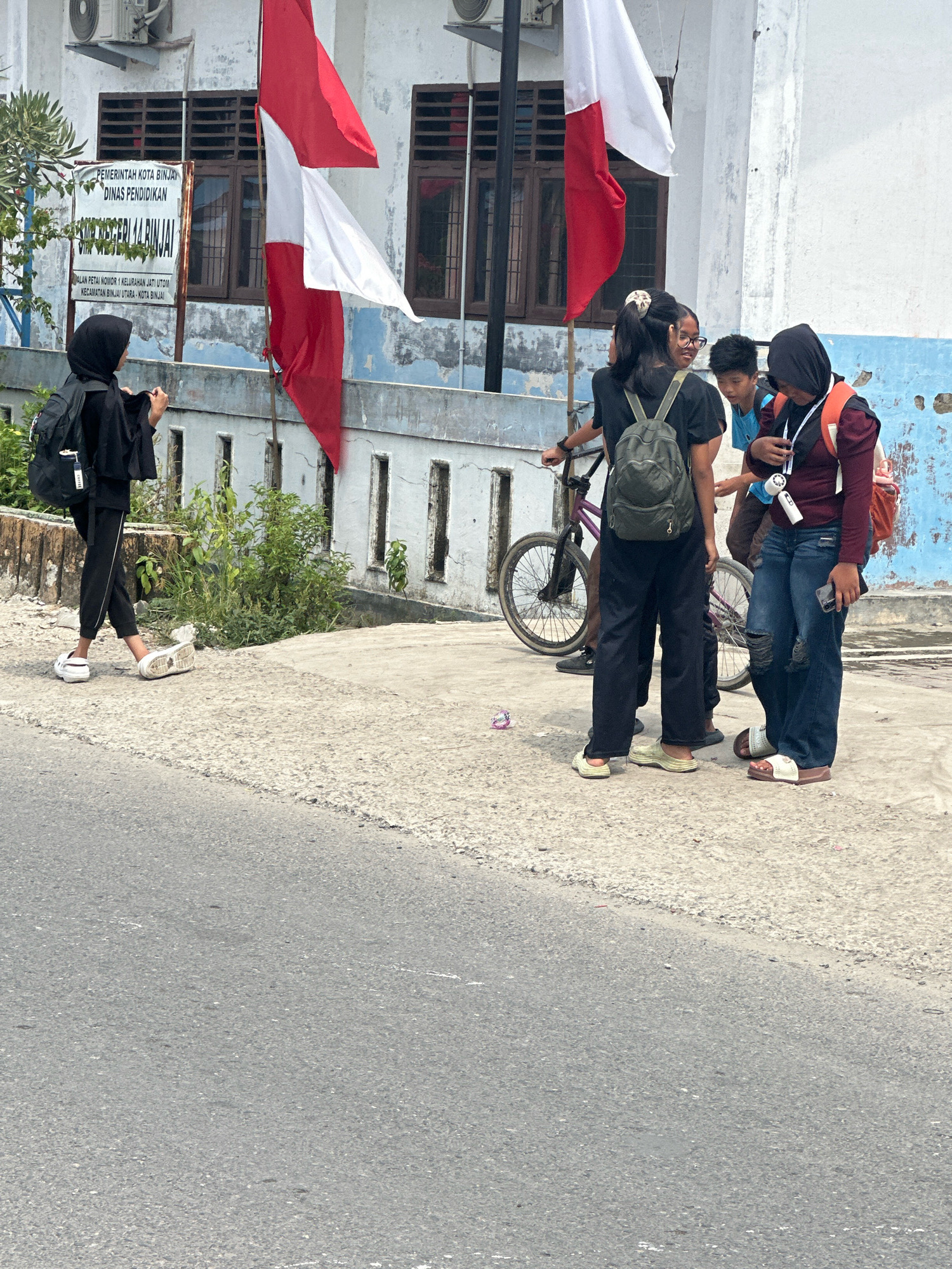 The image shows a group of four young people standing on a paved area next to a building in the city of Binjai, Indonesia. In the foreground, a person wearing a black hijab and black clothing, carrying a black backpack, is walking away from the camera towards the left. They are wearing white slide sandals. Behind them, the building has a light blue and white painted facade with several windows and two Indonesian flags (red and white) flying. A sign on the building reads "PEMERINTAH KOTA BINJAI DINAS PENDIDIKAN" and "SMA NEGERI 14 BINJAI." To the right of the person in the hijab, three other individuals are gathered, one with a green backpack, another in a blue shirt, and the third wearing a dark red top and jeans. A bicycle is also visible behind the group. The ground is a mix of concrete and asphalt. Lush green vegetation is visible to the left of the building.