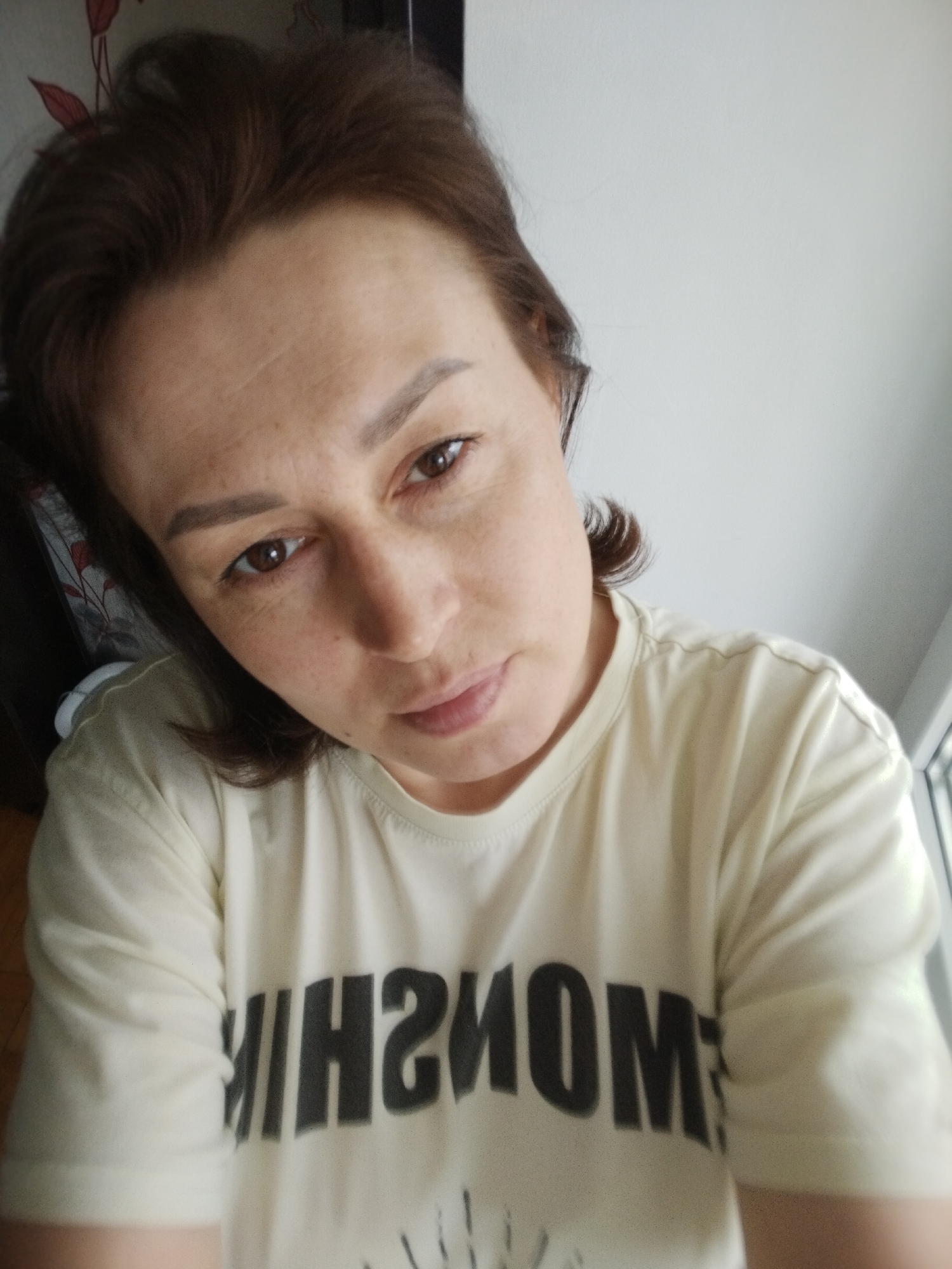 A woman with brown hair is seen in a close-up, slightly angled selfie. She has fair skin and brown eyes, and she is wearing a light-colored t-shirt with a dark, possibly mirrored, word printed on it. The background includes a white wall and a window. There are no discernible indicators to confirm the location as the city of Chaikovsky, Russia.