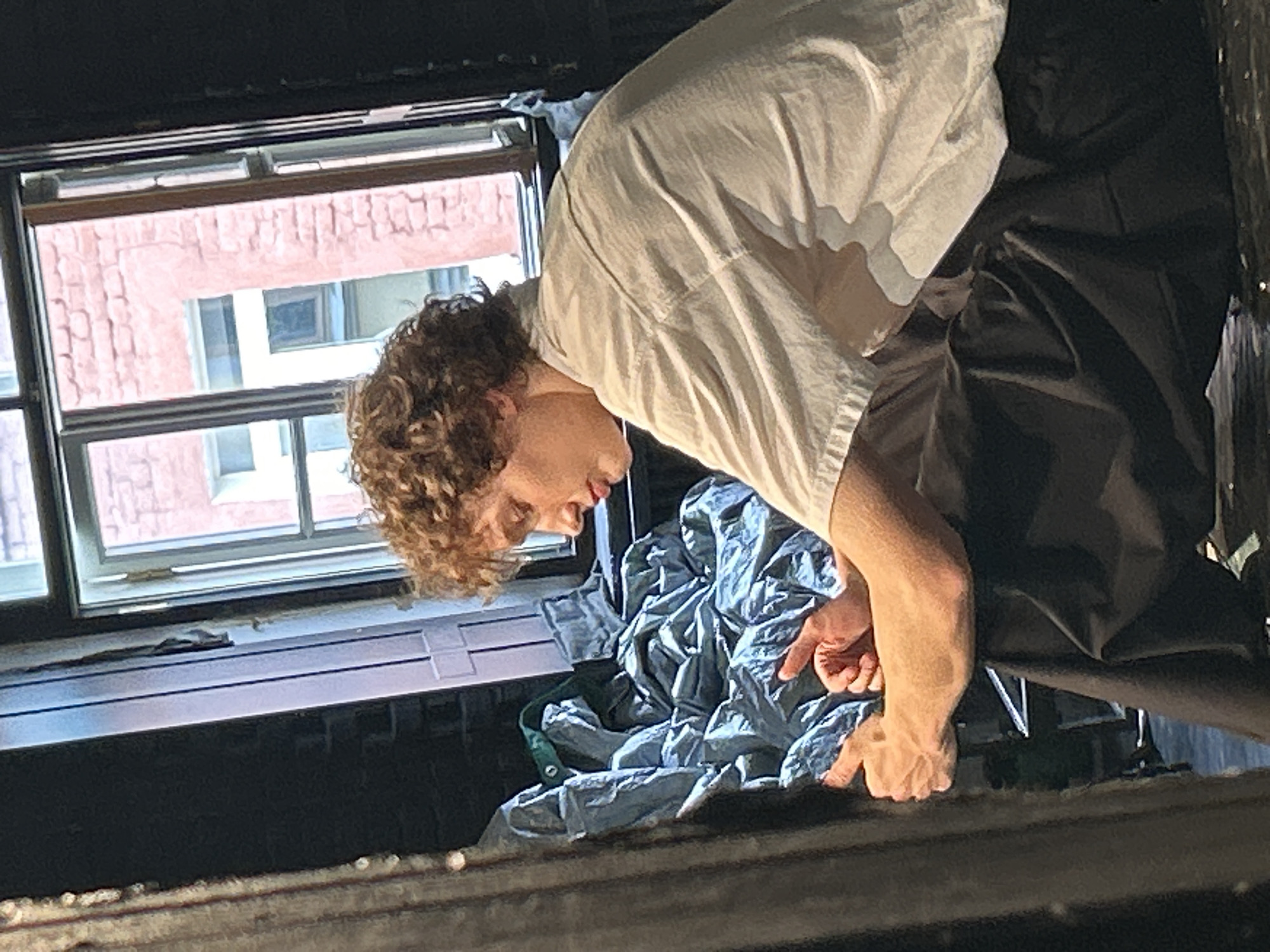 The image shows a person with curly brown hair leaning over and looking down. They are wearing a light-colored t-shirt and dark pants. Their arms are bent at the elbows, and their hands are visible in the foreground, with one hand appearing to be pointing or reaching towards something. The person's face is partially obscured as they are looking down and away from the camera.
Behind the person, a window is visible with a brick building outside. The window appears to be open or ajar. Below the window, there is a dark, metallic structure that seems to be a heating unit or ventilation system. A crumpled piece of light blue fabric, possibly a blanket or tarp, is visible between the person and the window.
The lighting suggests it might be daytime, possibly indoors with natural light coming from the window. The overall scene appears somewhat cluttered, with the fabric and the person's posture giving a sense of informal activity. There is no visible text in the image. The mood seems focused and possibly engaged in a task or observation.