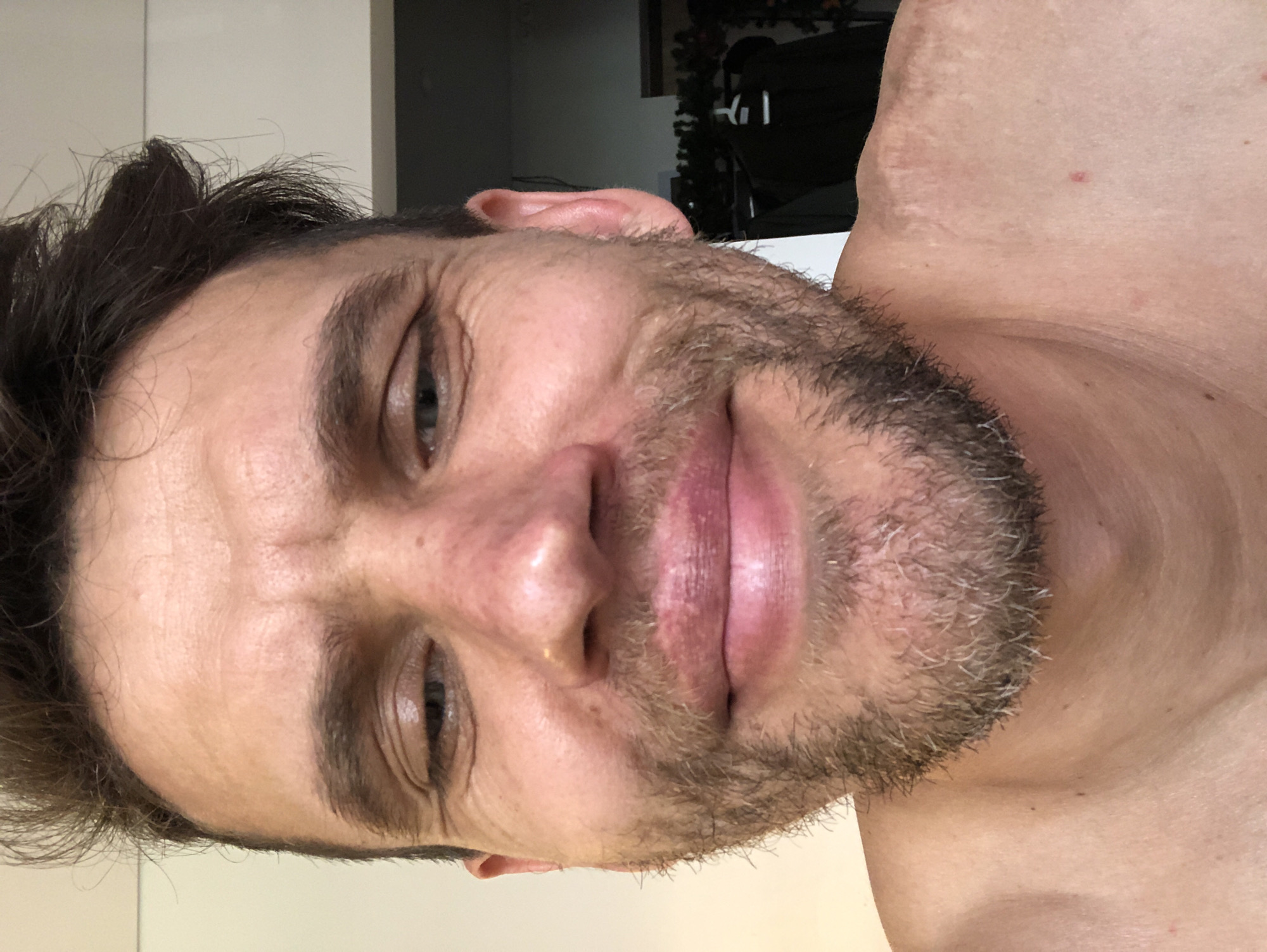 A close-up, low-angle selfie shows a fair-skinned man with dark, messy hair and a short beard. He is looking directly at the camera with his eyes partially closed and his lips slightly parted. His face is illuminated from the front. The background is out of focus, with hints of a wall and some clothing hanging on a rack. The overall impression is of a casual, intimate portrait. There is no discernible location in Saint Petersburg, Russia.