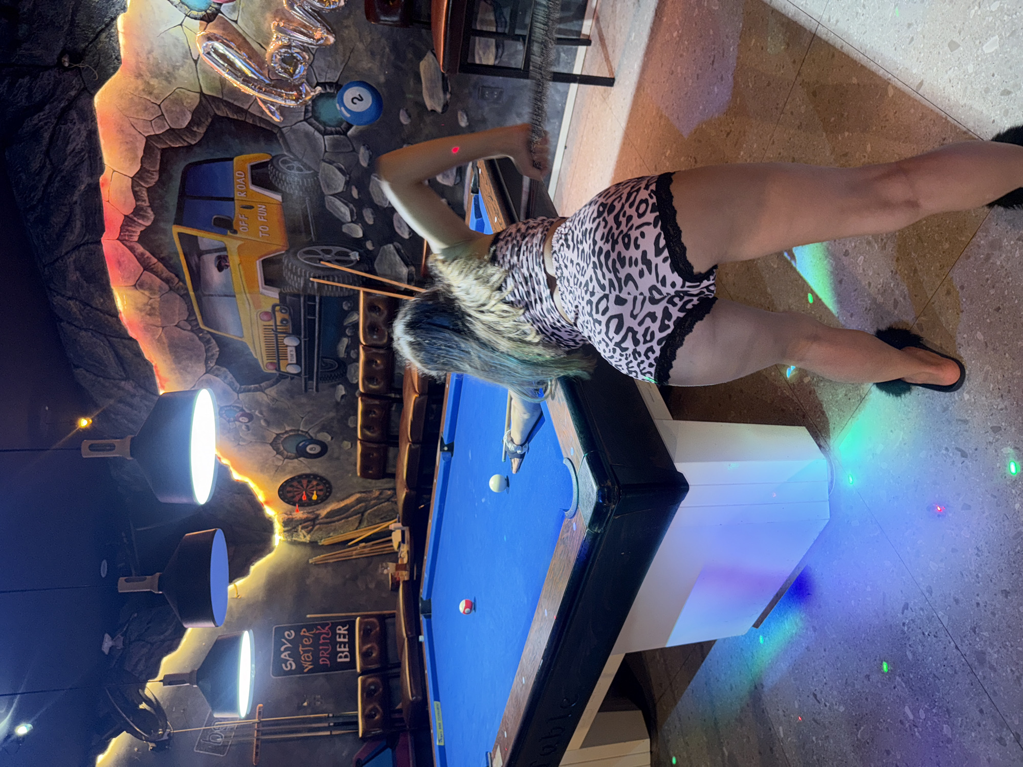 A woman in patterned shorts plays pool at a pool hall in Đà Nẵng, Vietnam. A pool table occupies the center of the frame. The woman is positioned on the right side of the pool table, in a stance that suggests she is about to strike the cue ball with a pool stick. Two balls are visible on the pool table. The pool hall features unique wall decorations, including a mural depicting a vehicle and text that says "OFF ROAD TO FUN." Additional wall decorations include images of pool balls and darts. In the background, a sign reads "SAVE WATER, DRINK BEER." Above, a series of three pendant lights illuminate the scene. The floor around the pool table is a tiled pattern and is illuminated with colored lights.