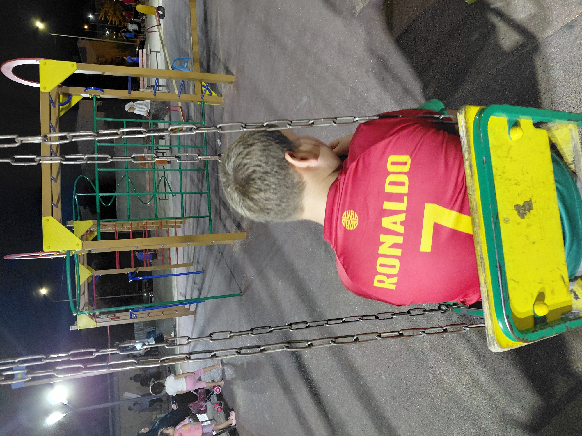 This is a night-time, medium-shot, eye-level photograph of a young boy sitting on a yellow and green swing set in a playground. The boy is viewed from behind, facing away from the camera. He has short, light brown hair and is wearing a red t-shirt with yellow lettering that reads "RONALDO" and the number "7". A yellow emblem is visible on the upper left side of the t-shirt. The swing seat is yellow with green trim and appears to be made of wood. Large metal chains suspend the swing from above.
In the background, a multi-colored playground structure with yellow, green, and red elements is visible. Several people are present in the playground, including children playing and adults observing. Some are standing near the playground equipment, while others are seated on benches or in the distance. The ground of the playground is paved with asphalt. Streetlights and building lights cast illumination on the scene, creating shadows. The location is identified as a playground in the city of Aktau, Kazakhstan.
