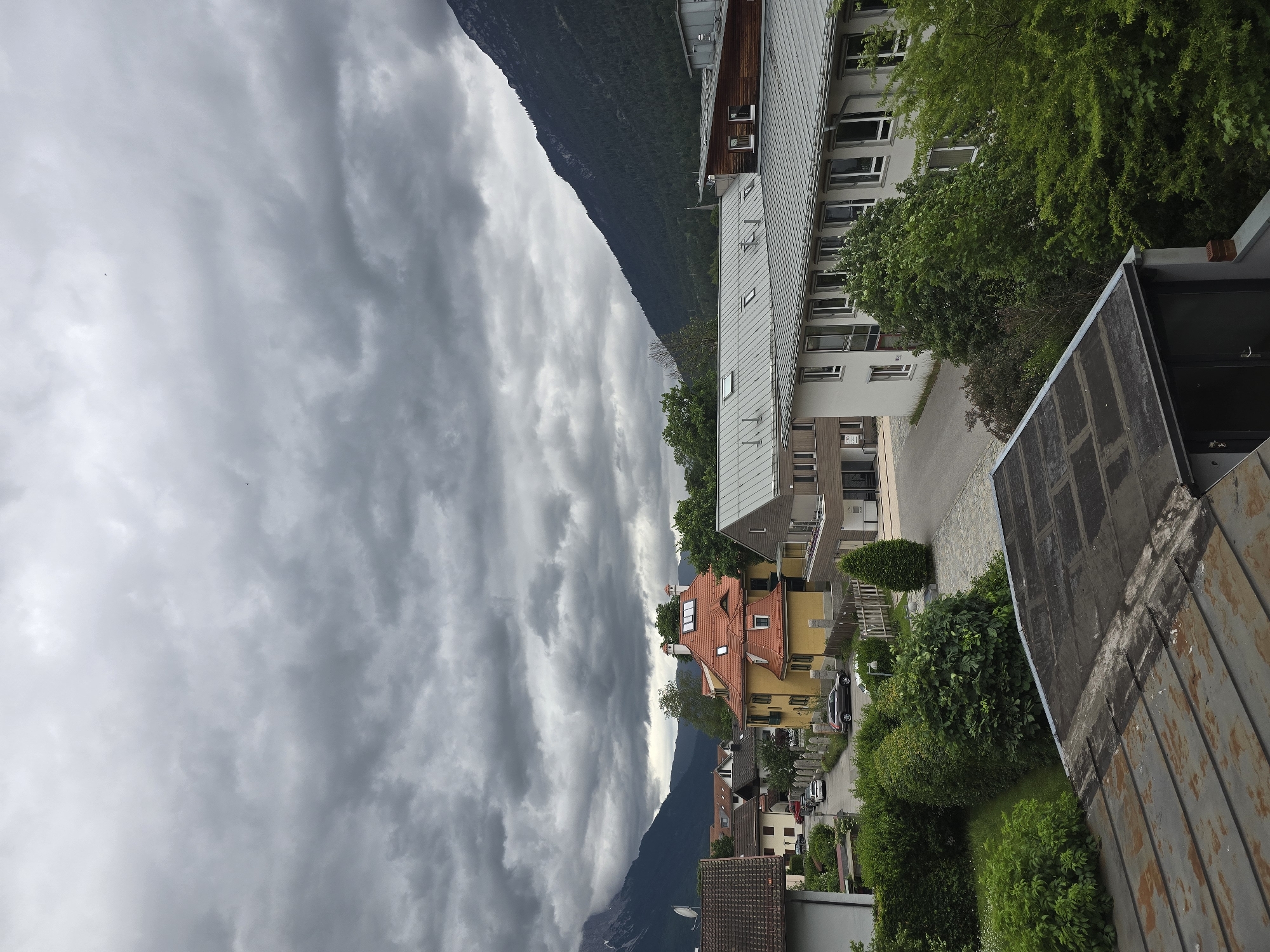This media file presents a panoramic view of a town nestled within a mountainous landscape, identified as Garmisch-Partenkirchen, Germany. The image is captured on an overcast day, with the sky dominated by dramatic, heavy grey clouds that suggest either an impending storm or recent rainfall.
In the foreground, on the right side of the frame, a textured, weathered metal roof structure with visible rust is partially visible. The midground features a cluster of residential buildings, showcasing various architectural styles. A prominent bright yellow house with a steep, red-tiled roof stands out among other structures, which typically have lighter-colored facades and grey roofs. Lush green trees and well-maintained bushes are scattered throughout the town, enhancing the verdant setting. A street or driveway is visible, with a few parked cars suggesting human presence, though no people are seen.
The background is dominated by majestic mountains. A large, dark green, forested mountain occupies the left and central parts of the frame, its slopes rising steeply towards the cloudy sky. Further mountains recede into the distance, partially obscured by the extensive cloud cover.
The lighting is diffused due to the thick clouds, creating a subdued but atmospheric scene. No specific activities or events are depicted, presenting a static snapshot of the town and its natural surroundings. There is no visible text in the image.