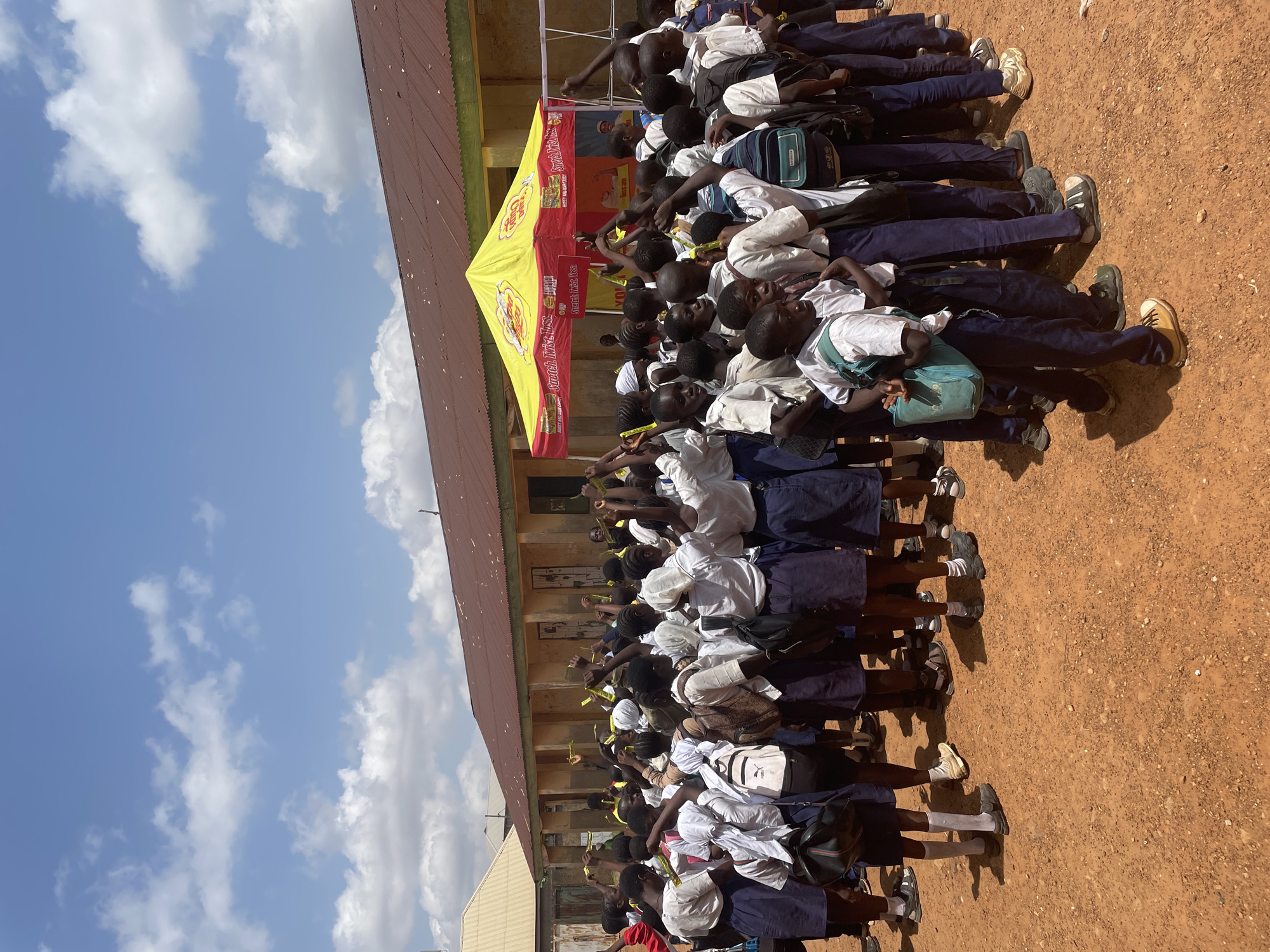 A group of schoolchildren are gathered outside a building, some standing and some sitting. They appear to be in a school setting, possibly during an event. The children are wearing school uniforms, consisting of white shirts and navy blue skirts or trousers. Many of them have backpacks. A yellow and red tent with visible text is set up behind them, suggesting some kind of promotional activity or outdoor event. The ground is reddish-brown earth, and the sky above is blue with scattered white clouds, indicating it's daytime and likely fair weather. The children are mostly looking in the same direction, and some are holding up yellow items, possibly promotional flyers or giveaways. The overall mood seems to be one of engagement and participation. The text on the tent reads: "Stretch. Twist. Toss. & Toss".