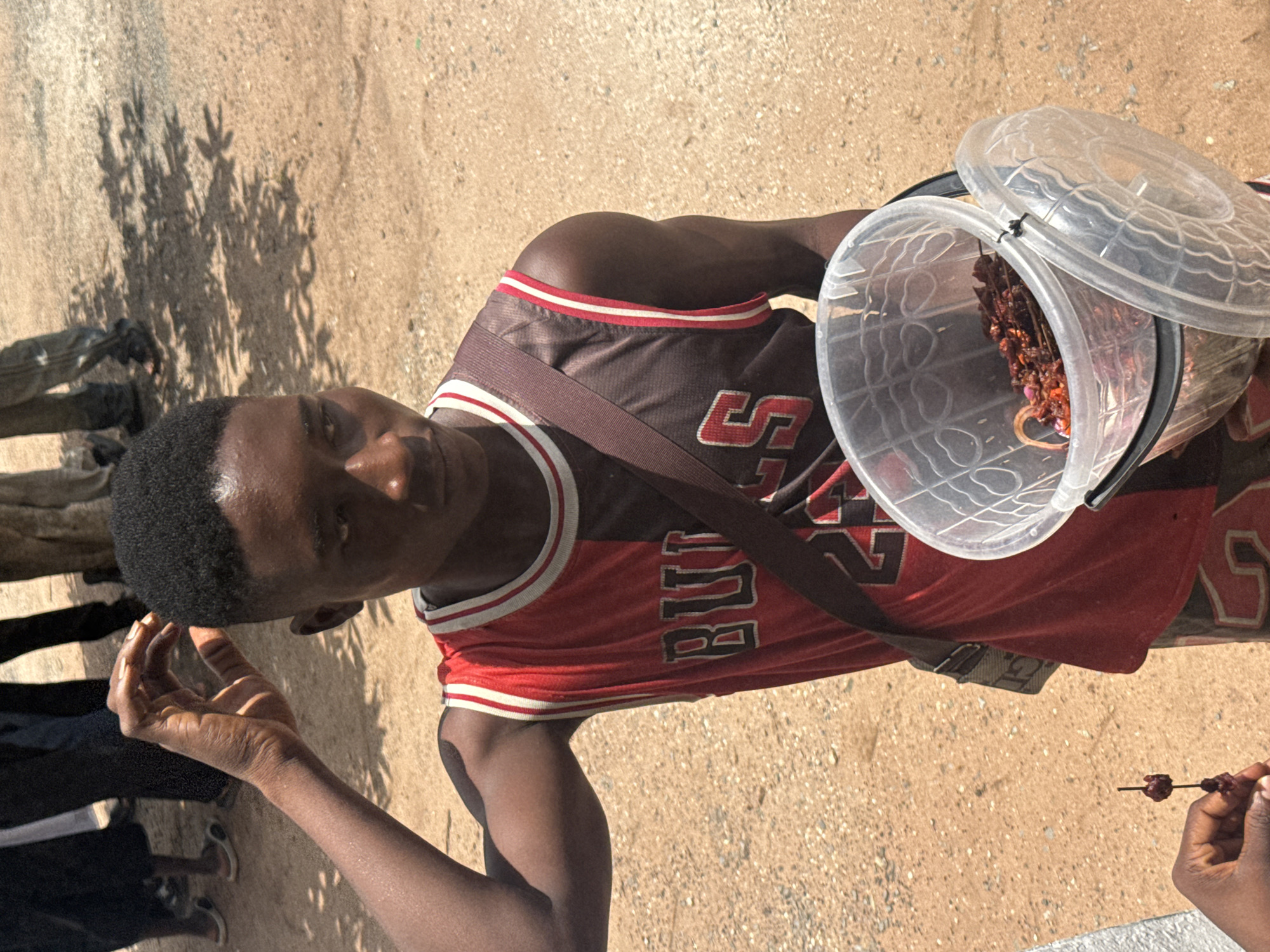 A young Black male wearing a red and brown sleeveless basketball jersey with the word "BULLS" and the number "23" is standing in a dusty outdoor environment. He has his right hand raised to his head, scratching his hair. He is wearing a dark strap across his chest and holding a clear plastic bucket with a lid, which contains dried red items, possibly food. In the lower left corner, a hand is holding a small dark object on a stick. In the background, several people are standing, their legs and lower bodies visible, with some wearing dark clothing and others in lighter-colored pants. The ground is covered in light brown sand and small stones, with shadows from trees cast across it. The overall scene suggests a market or street setting in Ihiagwa, Nigeria, where the individual may be selling goods.