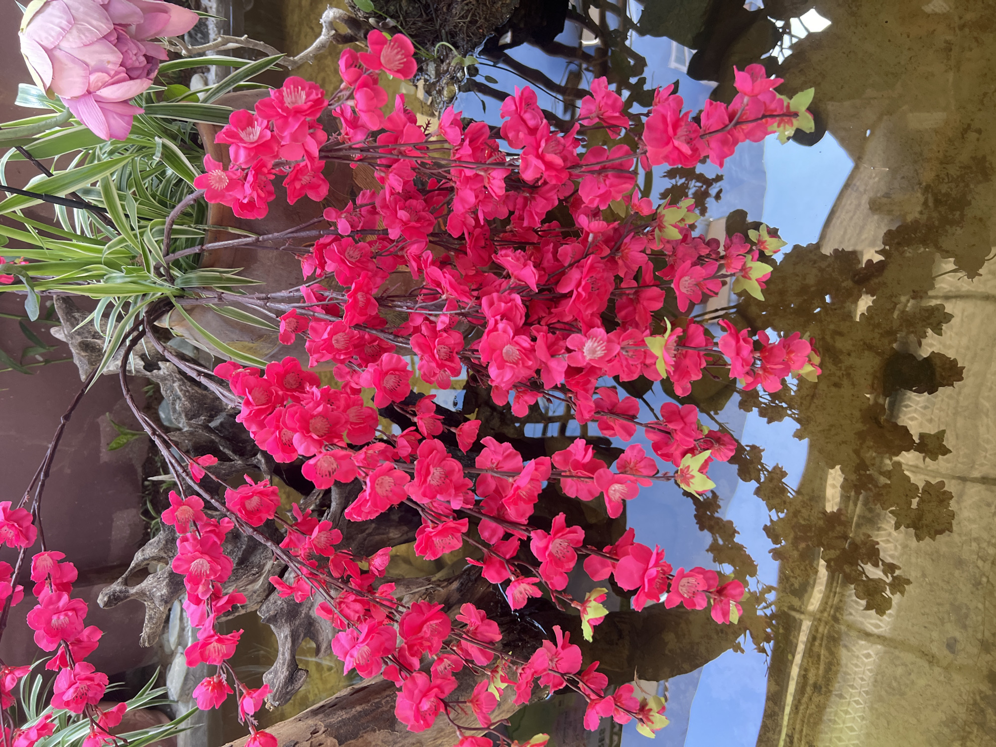 The image shows a close-up of bright pink artificial cherry blossoms hanging over a body of water. The blossoms are clustered on thin branches and some have small, light green leaves. In the background, there are green plants with striped leaves and a partially unfurled pink lotus flower in a brown ceramic pot. The water below reflects the blossoms and surrounding elements, creating a blurred, abstract pattern. The scene appears to be an outdoor decorative arrangement, possibly in a garden or courtyard. The location provided, Lùng Tám Commune, Vietnam, suggests a rural or village setting, though the image itself does not contain specific visual cues to confirm this locale.