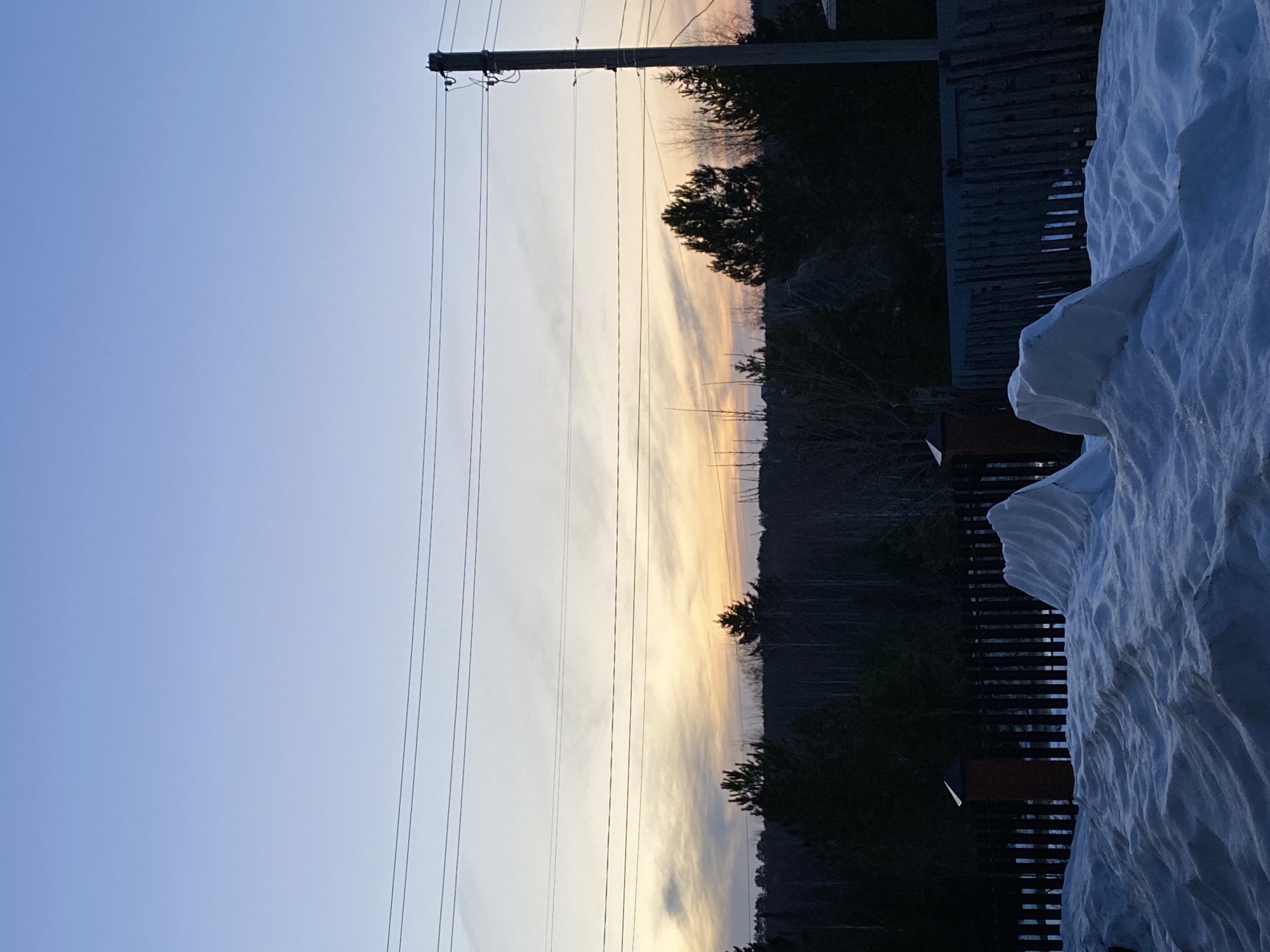 The image depicts an outdoor scene in Фоки, Russia during twilight. The sky transitions from blue to orange near the horizon, with utility poles and power lines visible in the foreground against the sky. Beyond the power lines are silhouetted trees and possibly a distant hill or mountain. To the right of the image is a wooden fence covered with a substantial accumulation of snow.