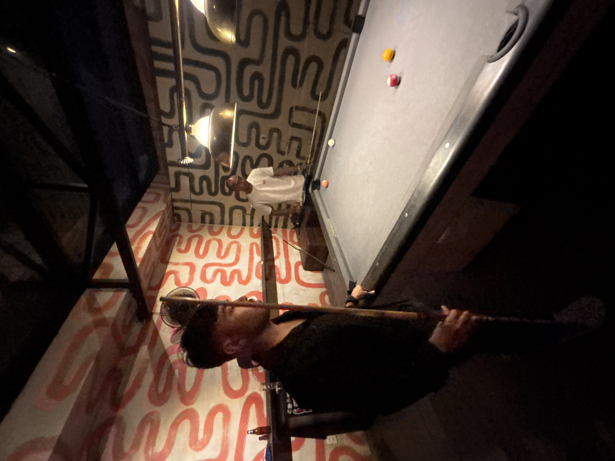 Two men are playing pool in a dimly lit room. The man on the left, facing away from the camera, is holding a pool cue. He is wearing a dark shirt and appears to be in motion. The man on the right, partially visible, is wearing a white t-shirt and is positioned behind the pool table, appearing to be about to take a shot. The pool table has several colored balls on it. The walls are decorated with a bold, abstract pattern in red and black. The room has exposed ceiling beams and industrial-style pendant lights. The setting suggests a bar or a game room. It is not possible to determine the specific location in Da Nang, Vietnam from the image.